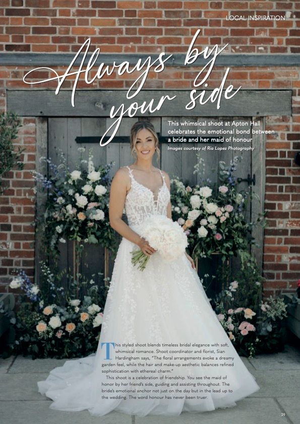 Bride in a lace wedding gown standing against a brick wall decorated with white floral arrangements.
