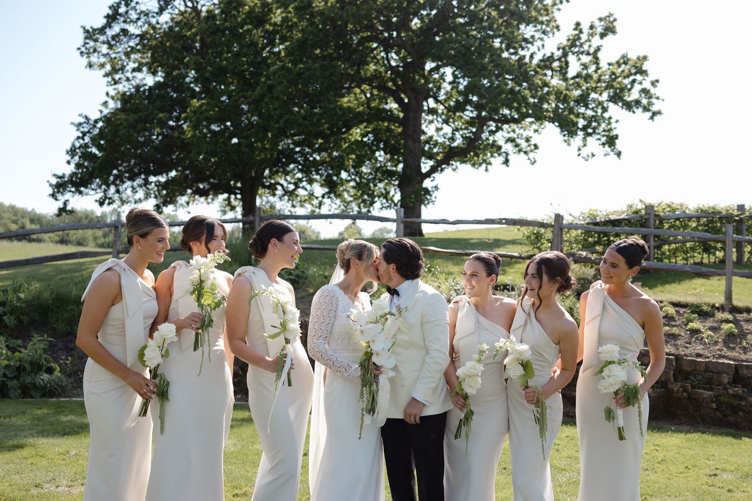Bridesmaids in white dresses holding bouquets stand outdoors as two brides share a kiss at their wedding.