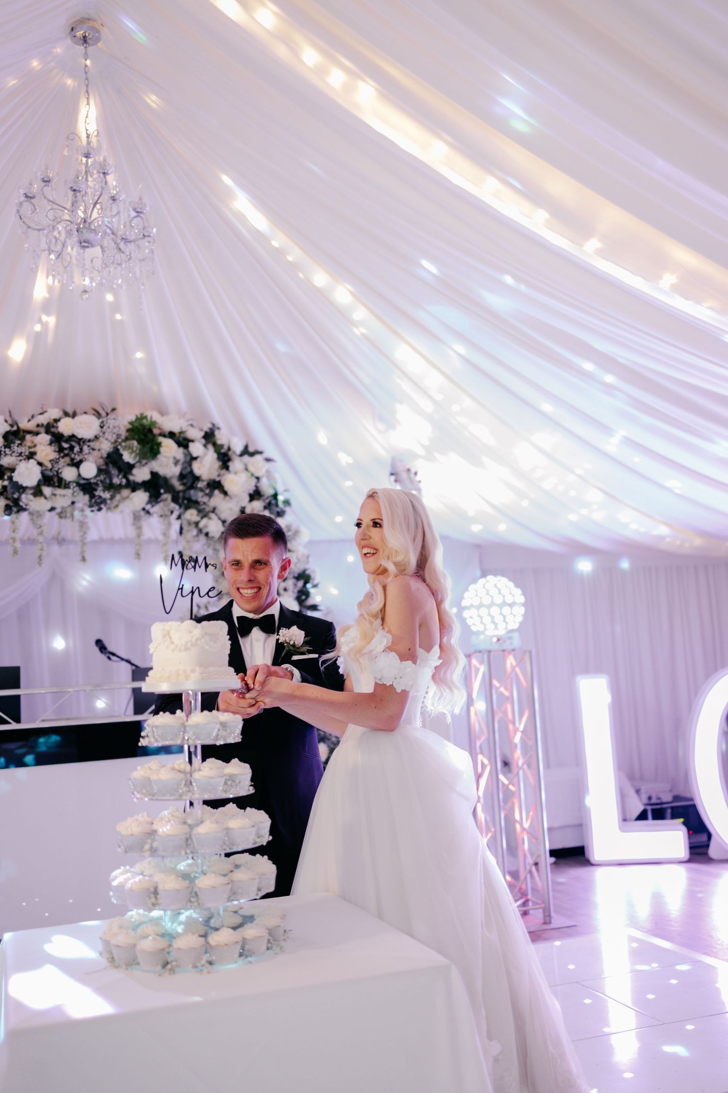 Wedding couple cutting cake at reception under white draped ceiling with fairy lights.