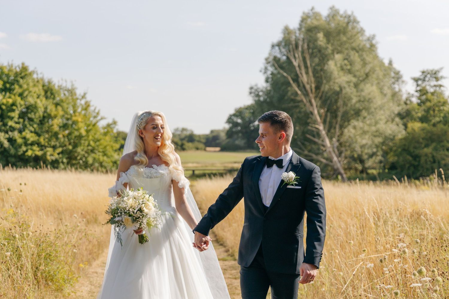 Romantic wedding portrait of couple walking through sun-lit summer meadow.