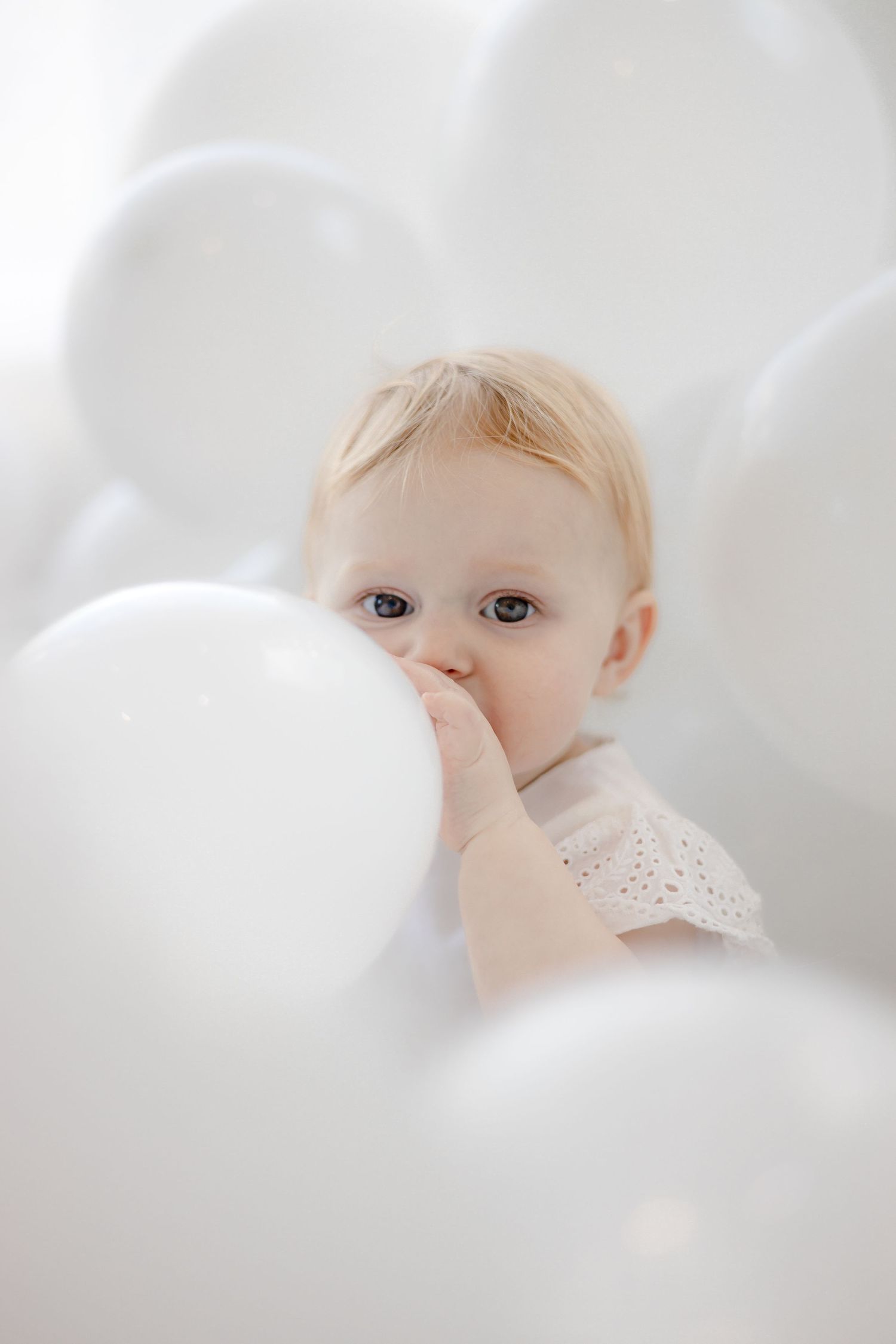 Baby peeks through white balloons in dreamy, ethereal portrait setting.