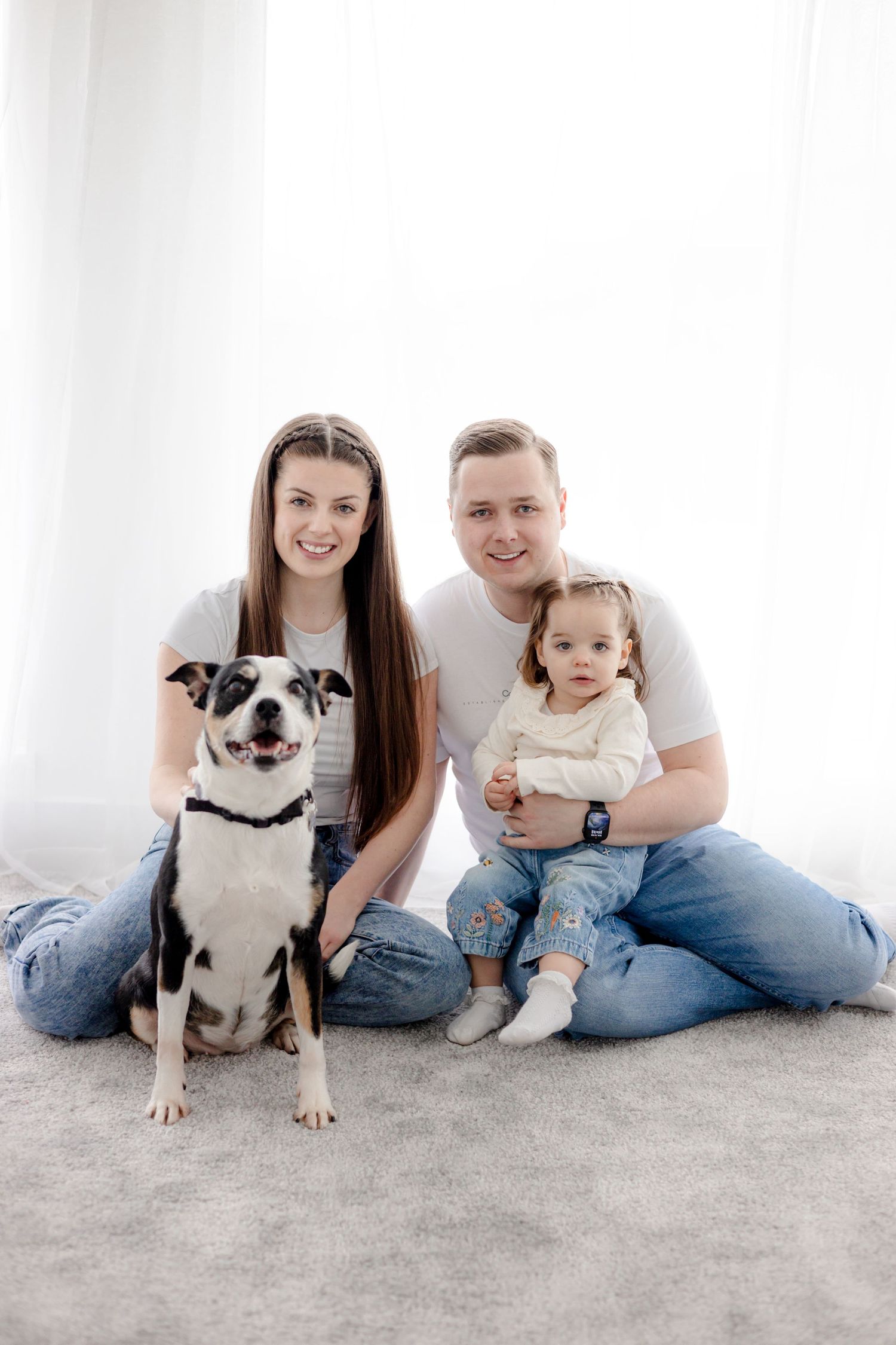 Happy family portrait with their black and white dog on light gray carpet.