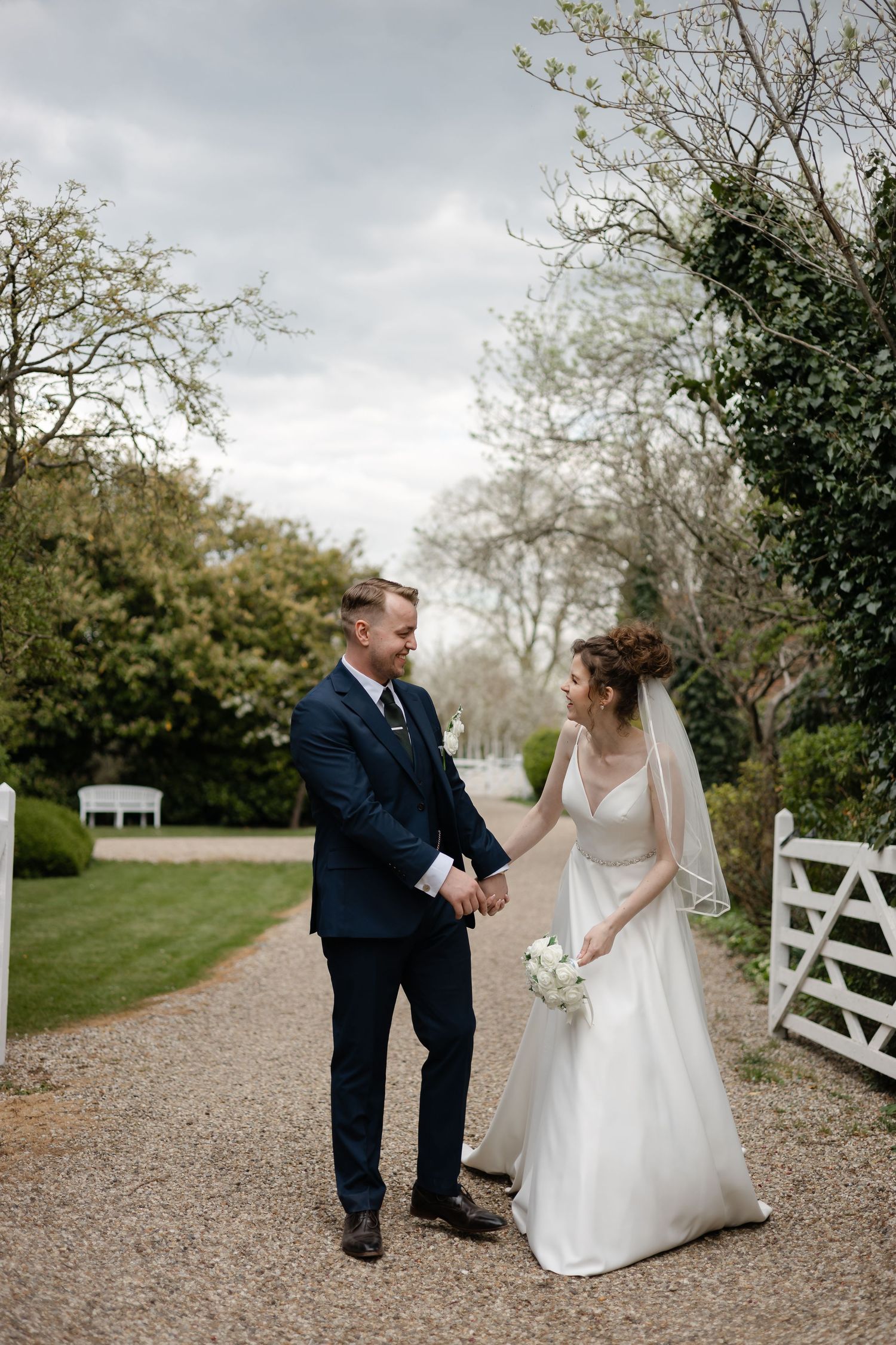 Couple shares a romantic moment on a gravel path near a white garden gate during their wedding day.