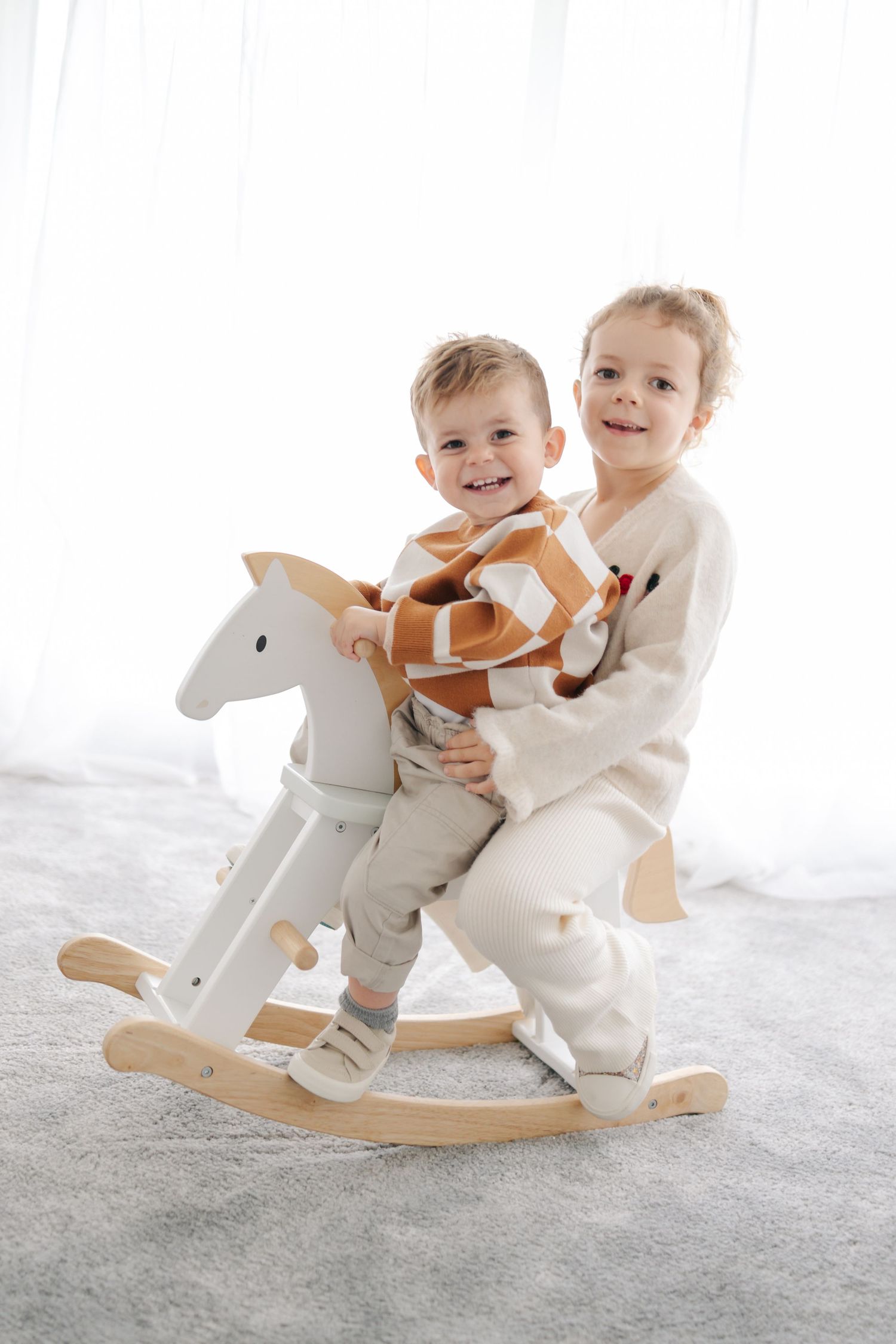 Two children in white outfits playing together on a wooden rocking horse against a light background.