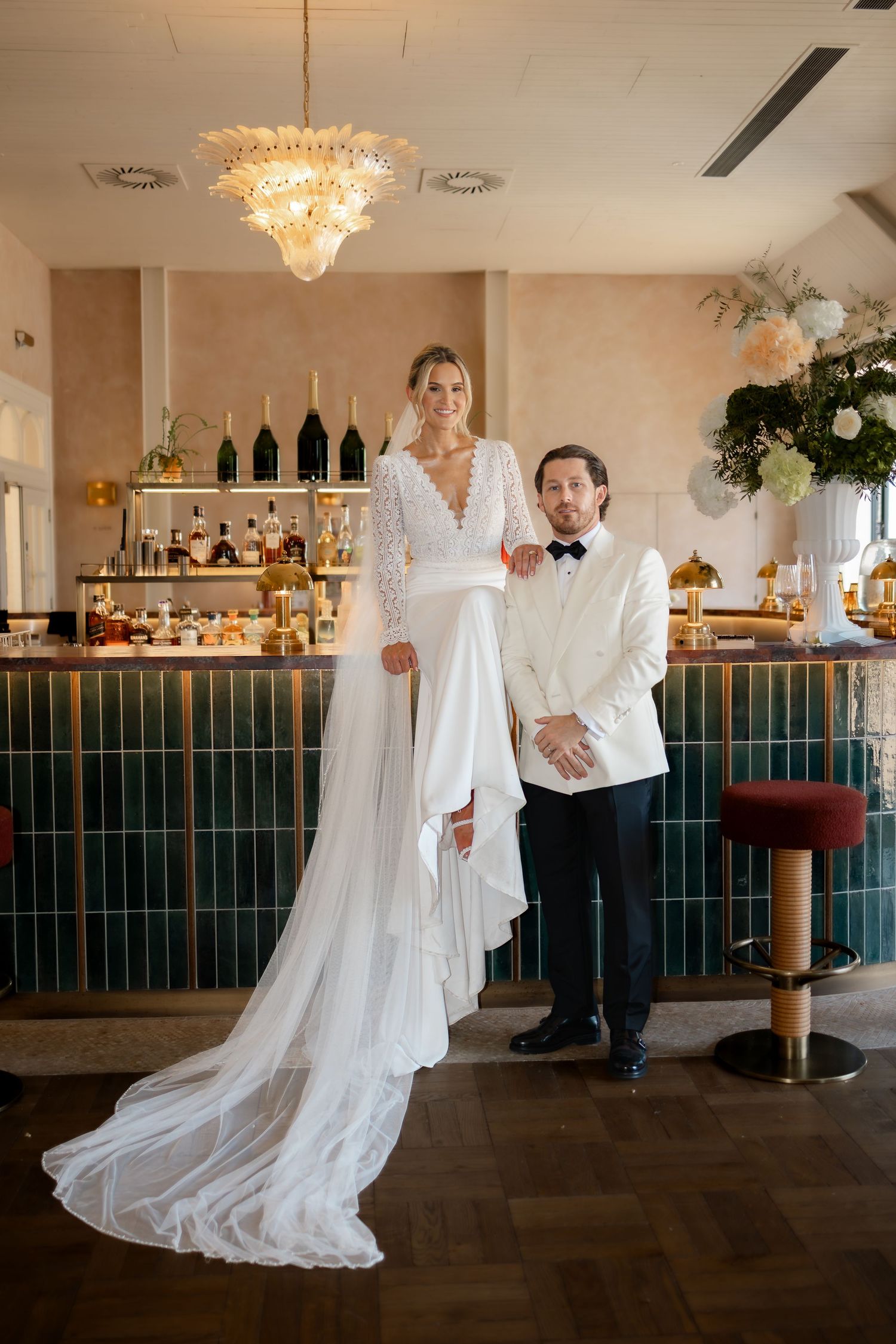 Elegant bride in long lace gown poses with groom in white jacket before a stylish green tiled bar.