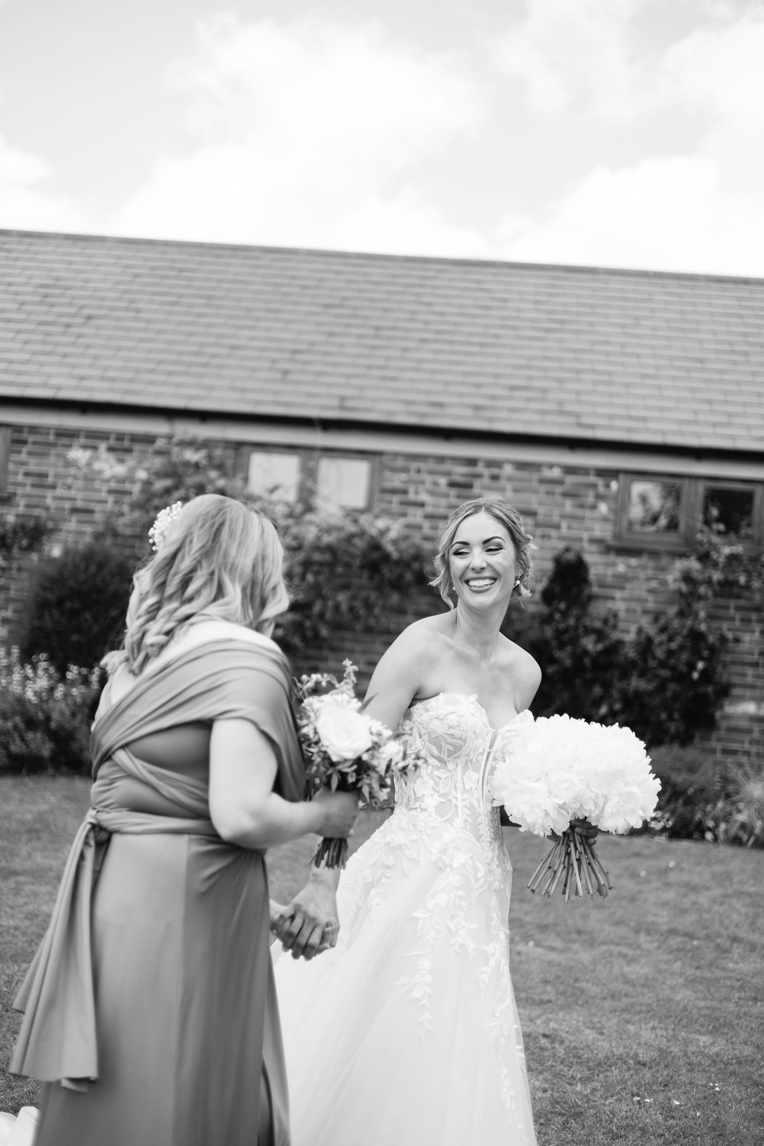 Bride in white lace gown shares joyful moment with bridesmaid in sage green dress on grassy lawn.