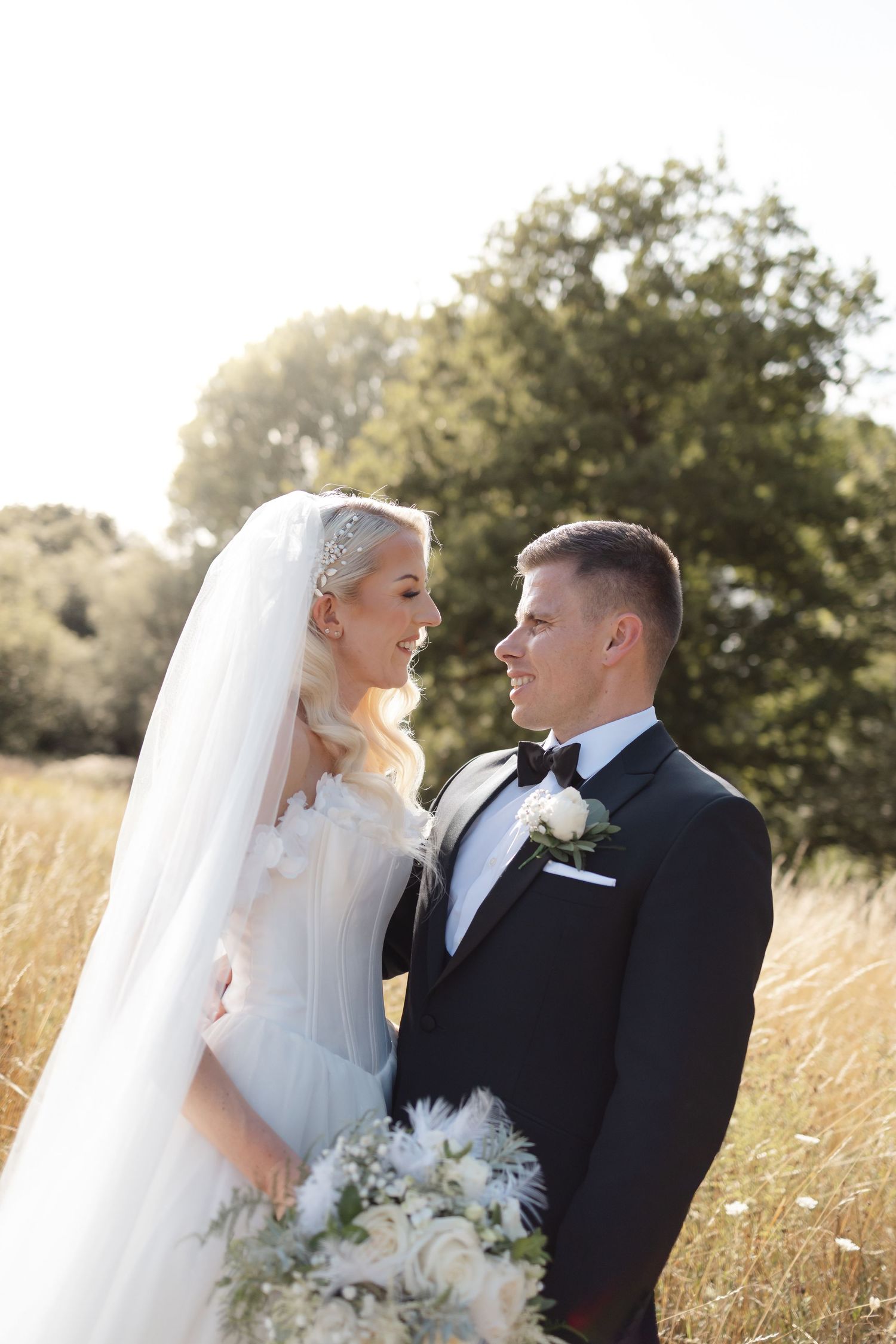 Elegant wedding couple poses in golden field with bride's veil flowing in natural light.