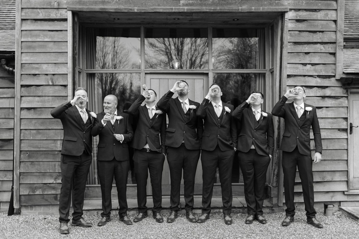 Groomsmen in black suits lined up against rustic wooden wall for wedding photo.