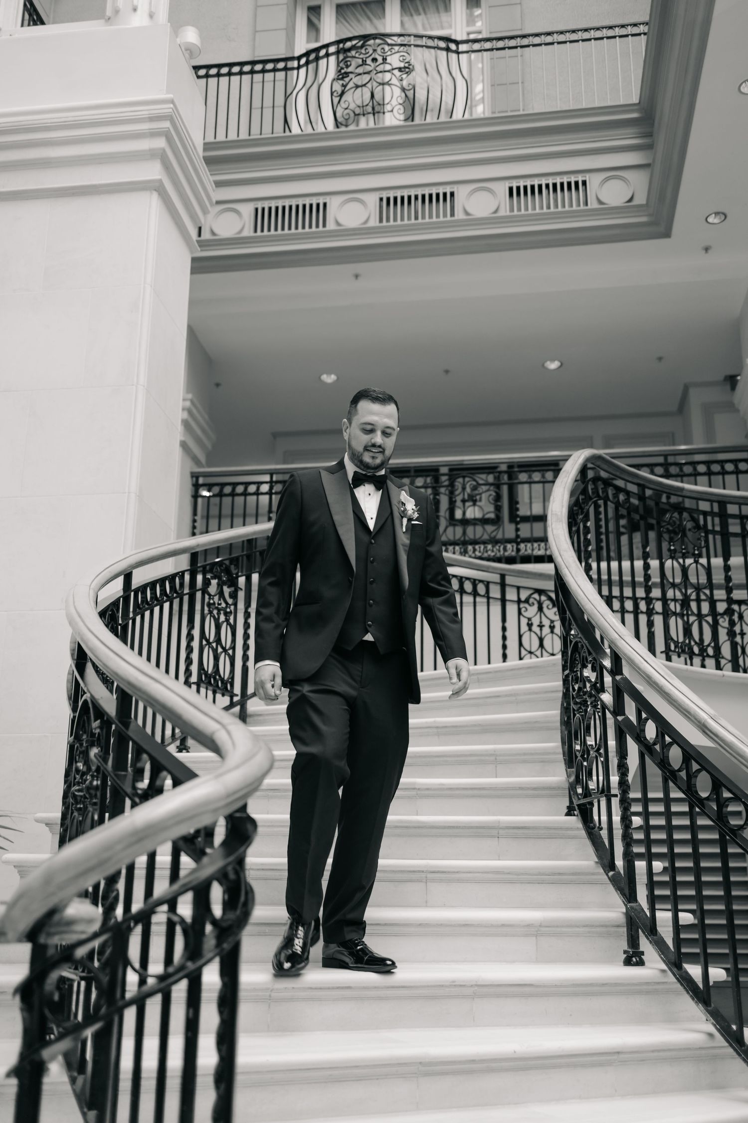 A groom in a black suit stands on an elegant white marble staircase with ornate black railings.