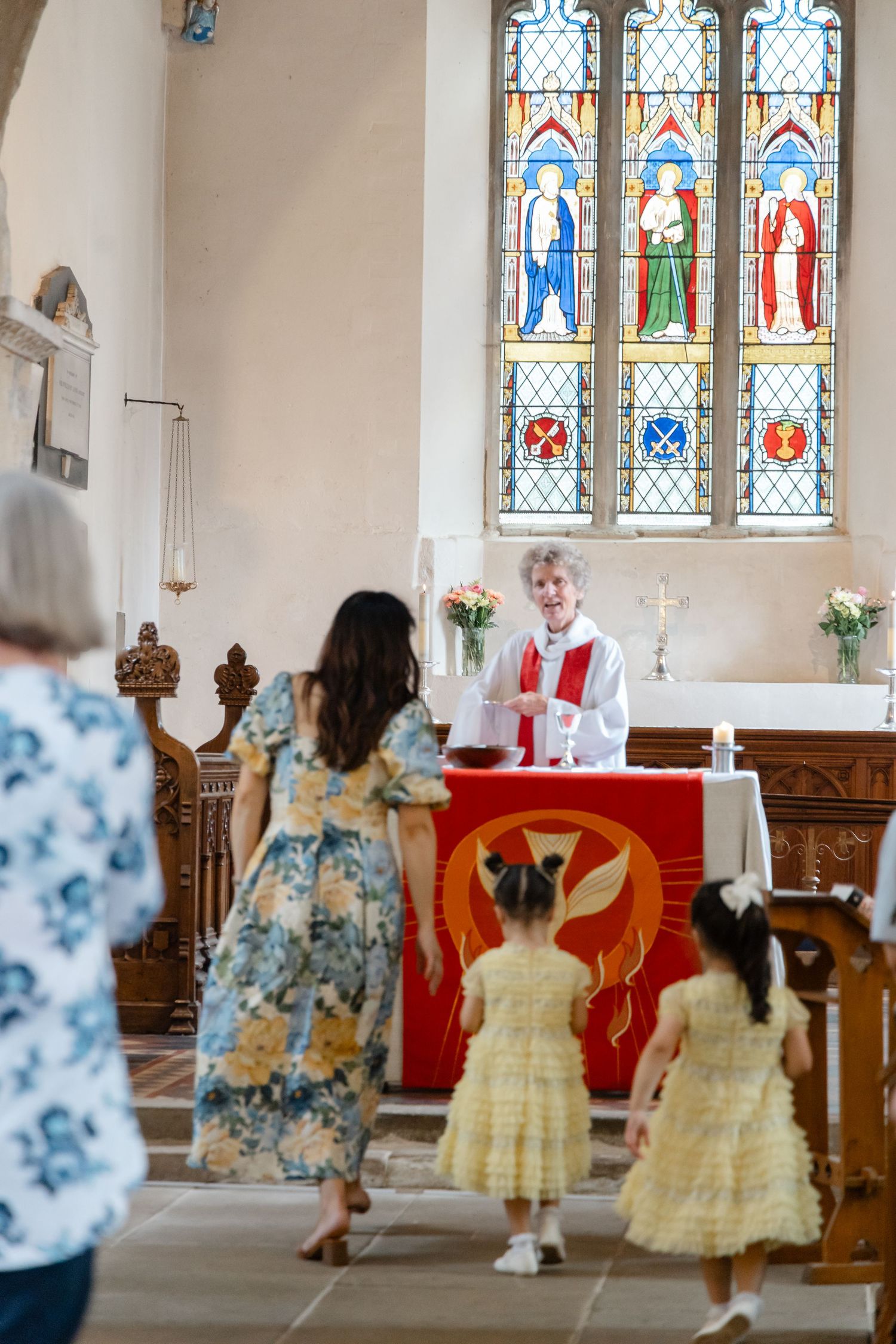 Children in floral dresses approach the altar during a church service with stained glass windows.