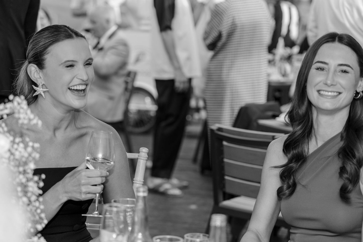 Two women laugh and smile while enjoying drinks at an outdoor social gathering.