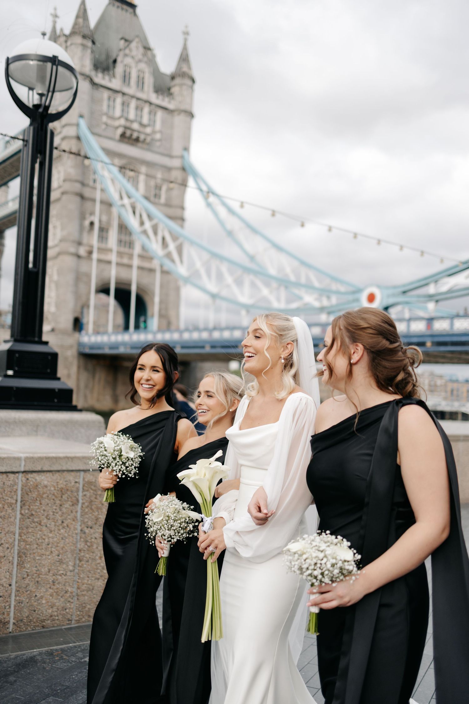 Bridesmaids in black dresses with white bouquets pose by Tower Bridge.