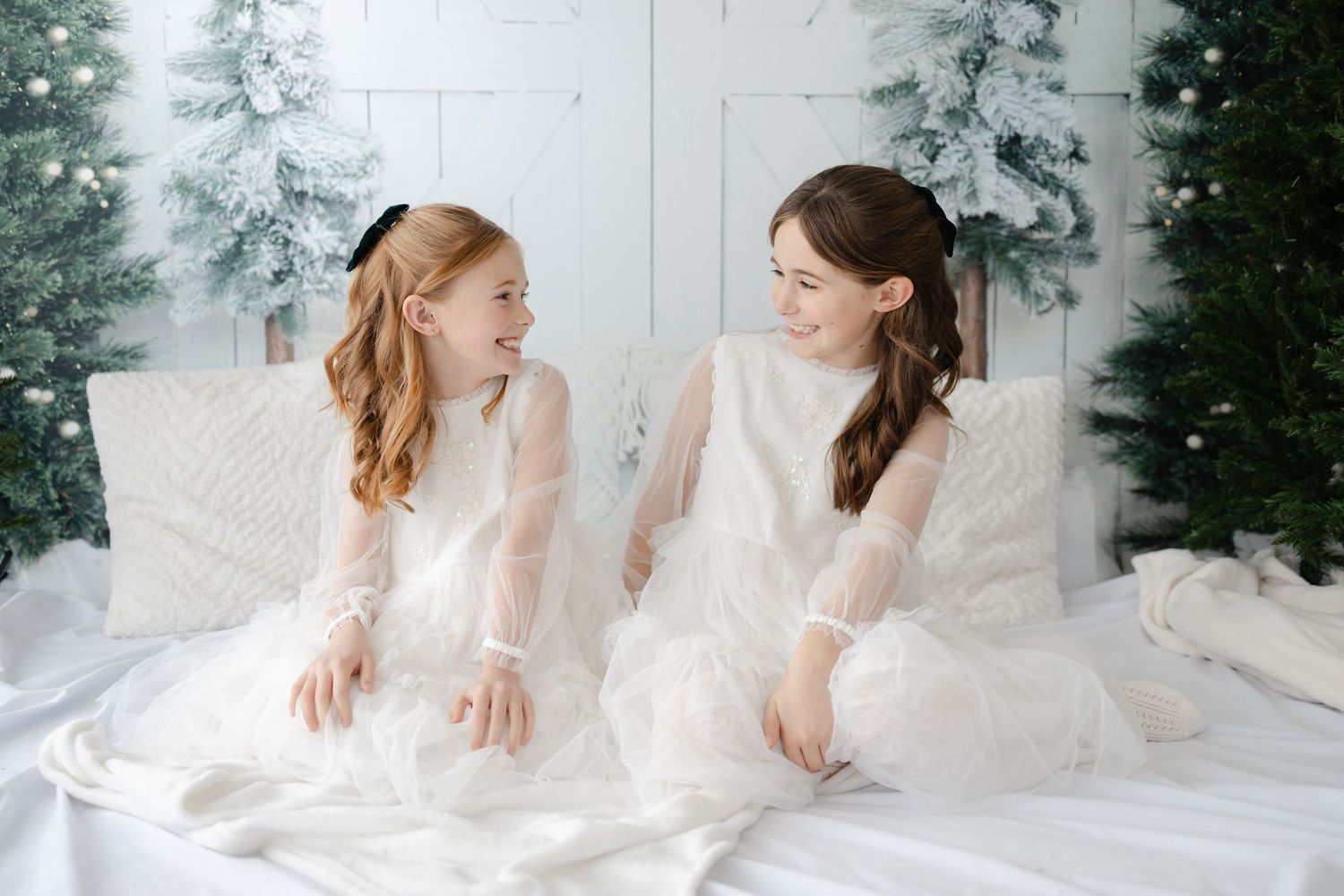 Two people in white dresses pose together on white bedding with winter decorations.