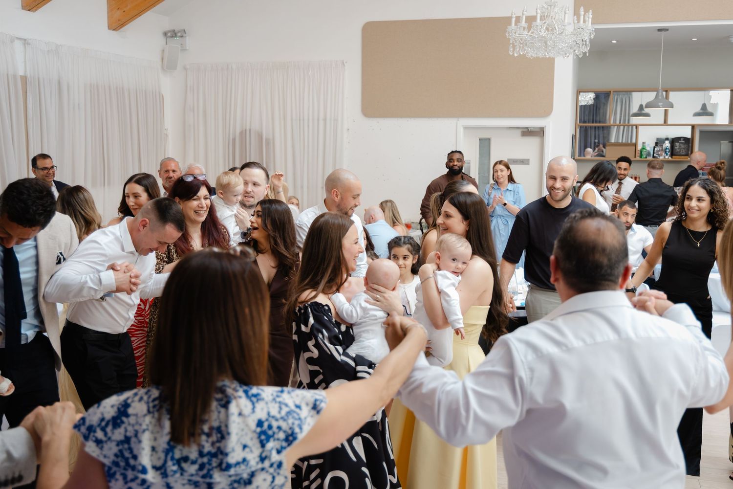 A lively crowd of guests dancing and celebrating in an elegant venue with white drapes.