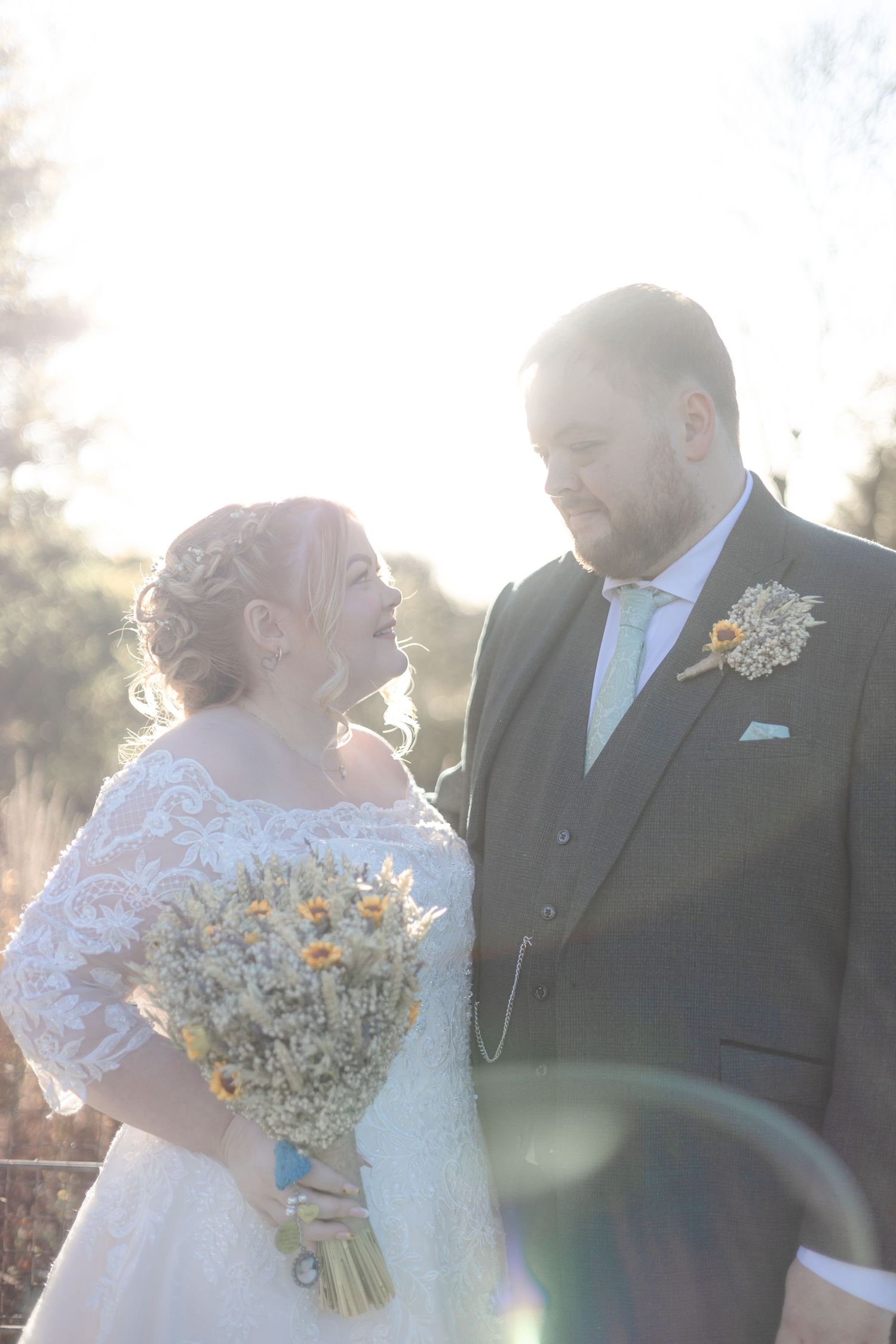Romantic wedding couple embraces in sunlit outdoor setting with rustic bouquet.