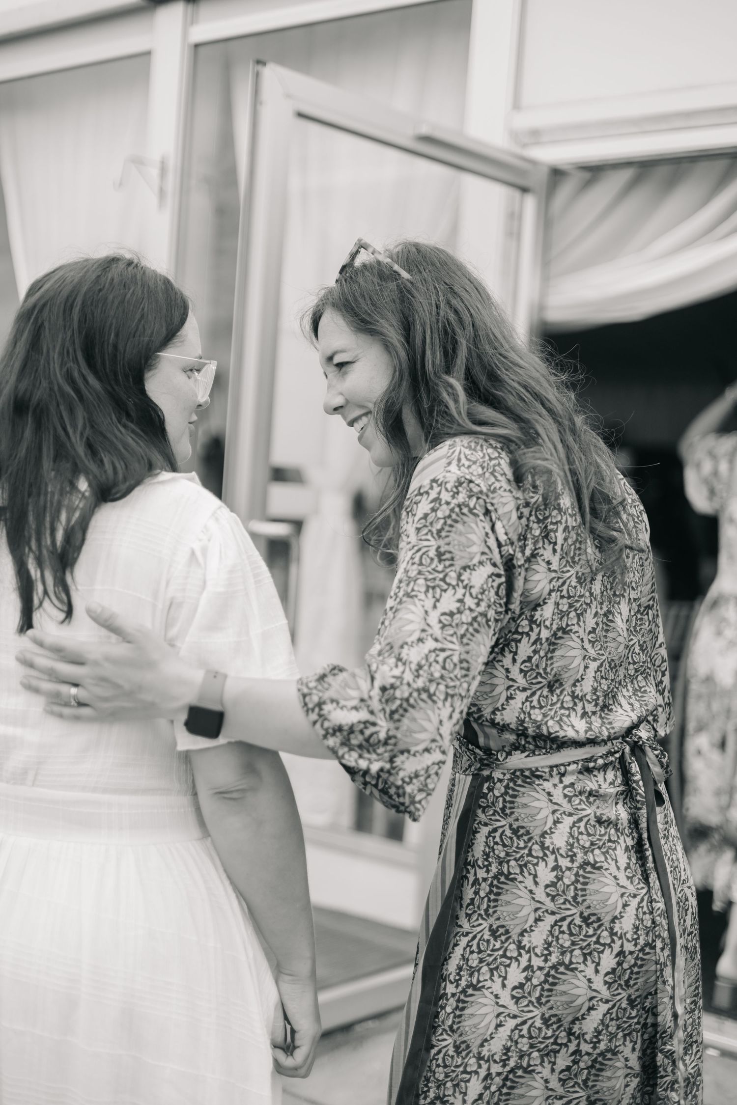Two friends sharing a joyful moment at an event, one wearing a white dress and another in a patterned dress.