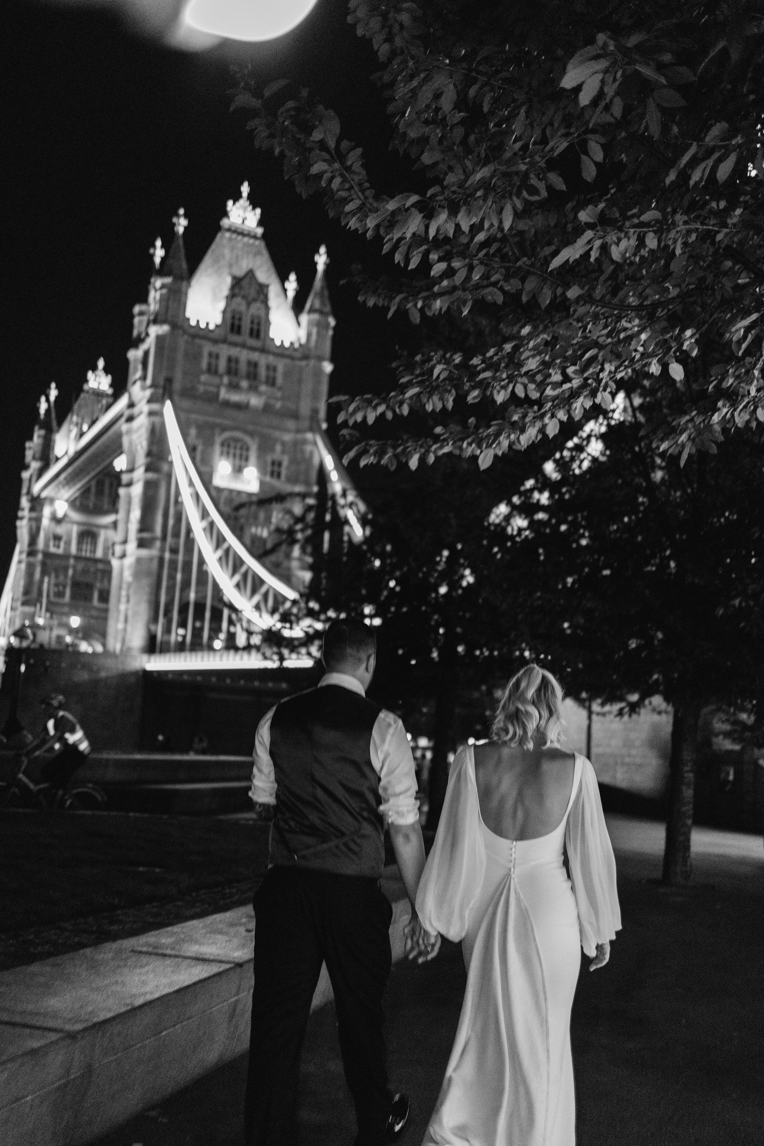 Couple walking at night with illuminated Tower Bridge in the background.