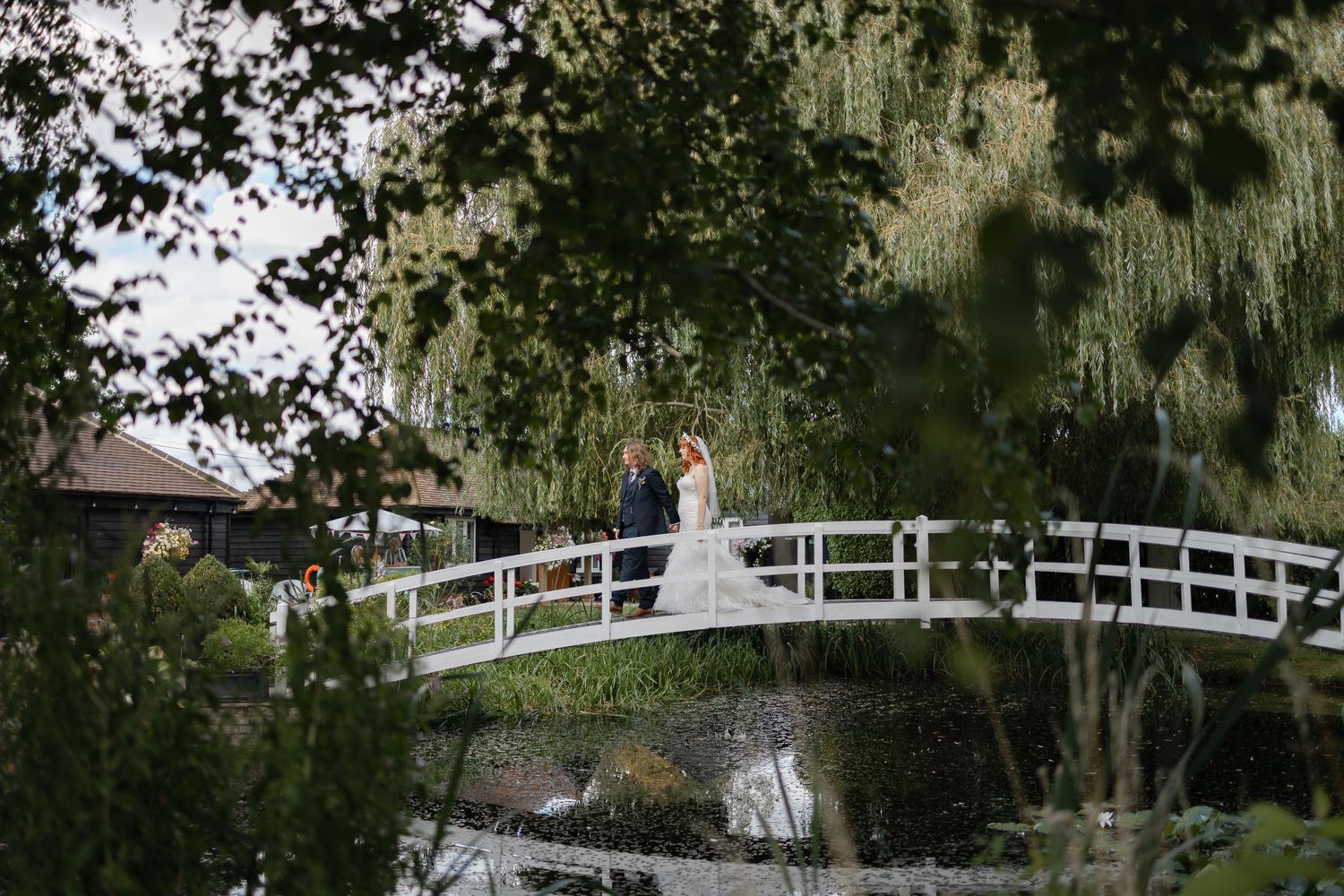 A wedding couple standing on a white wooden bridge surrounded by lush greenery.