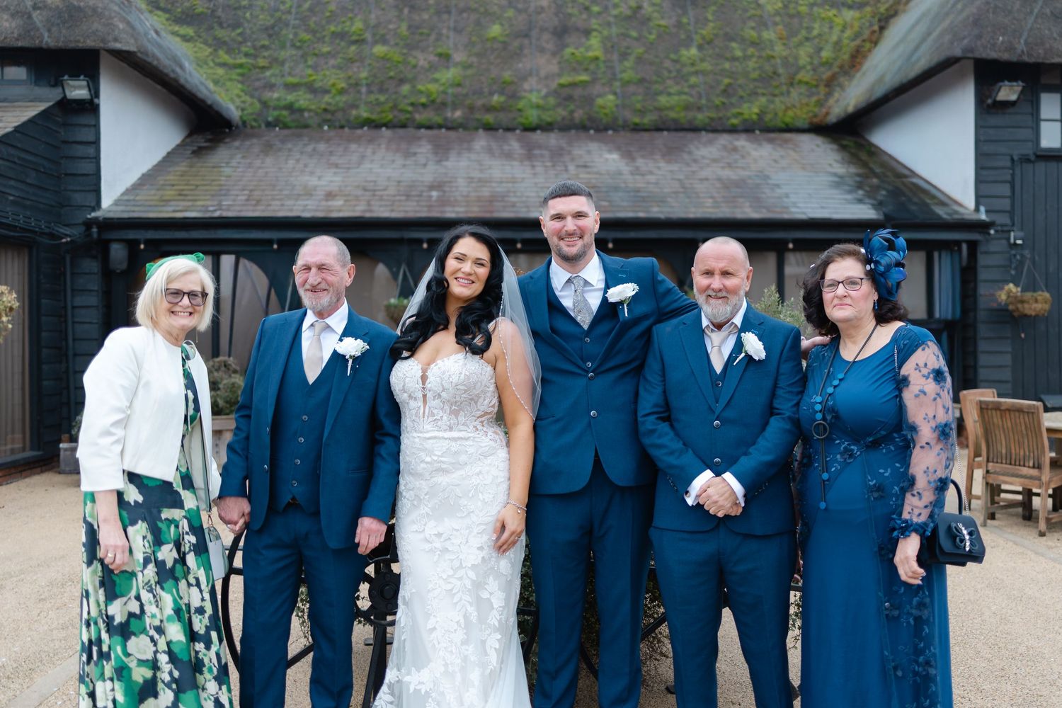 Wedding party in navy suits and light dresses posed outside a rustic building.