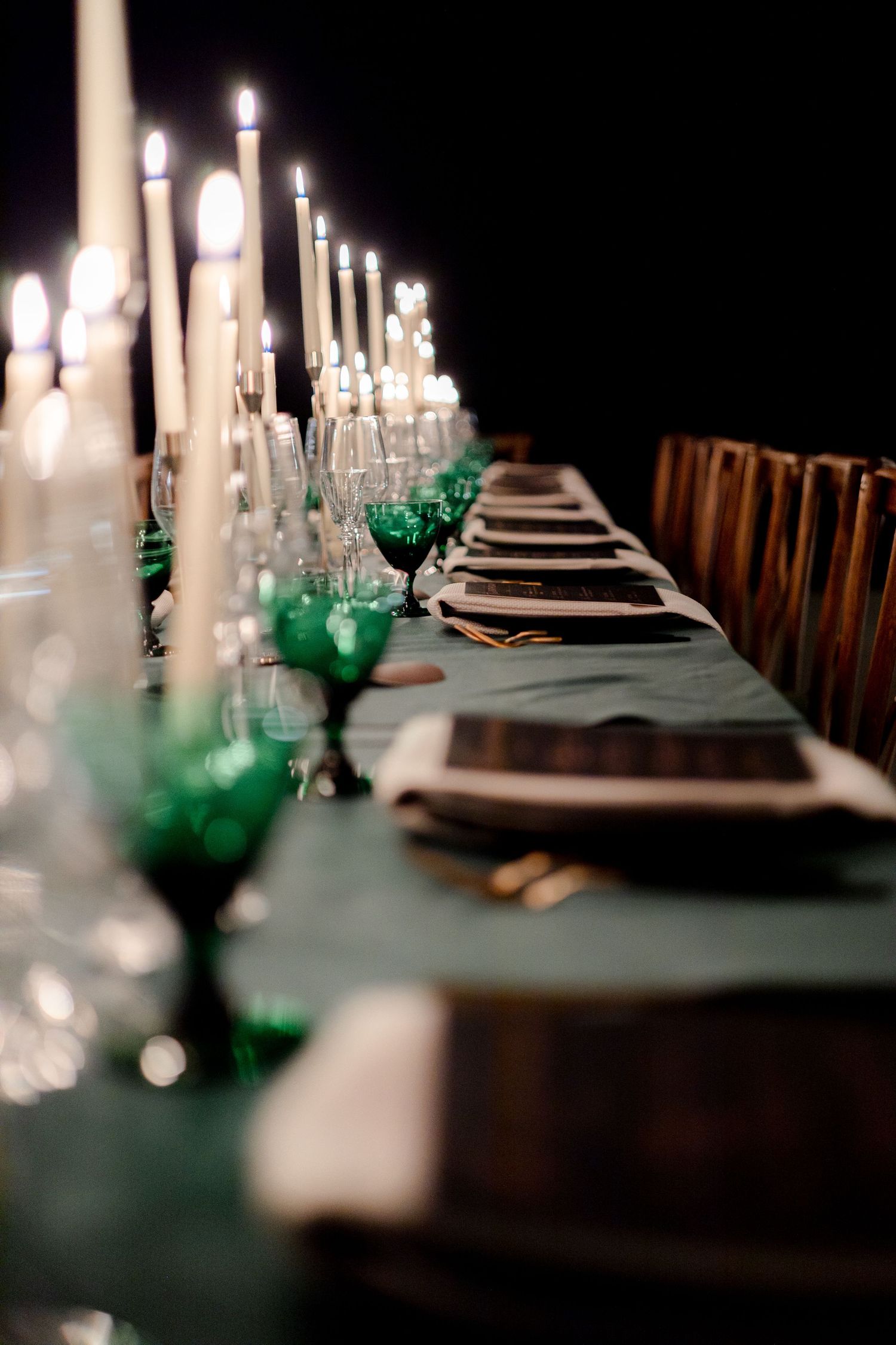 Elegant table setting with white candles and emerald green glassware against dark background.