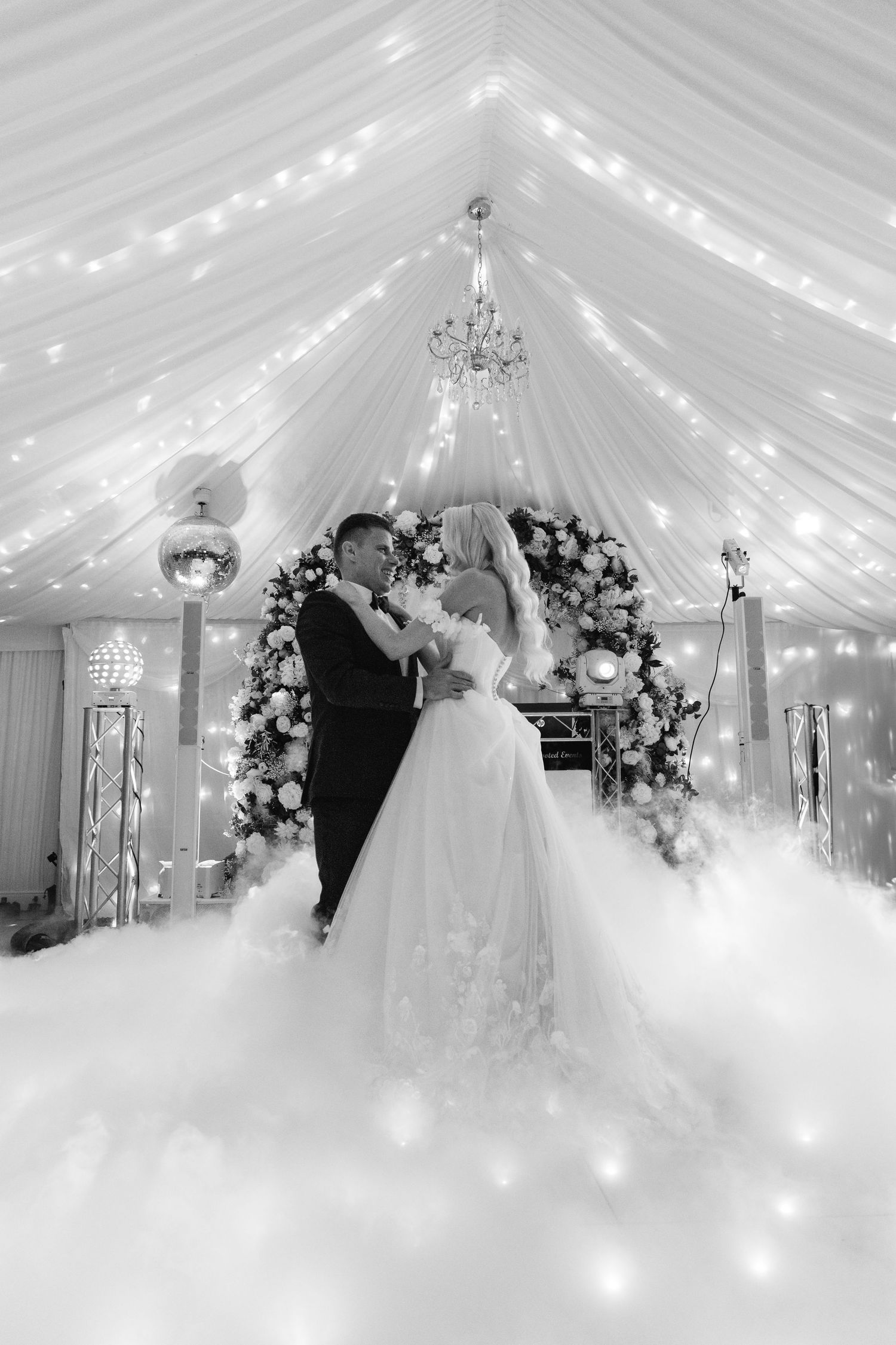 Romantic wedding reception under a white draped tent ceiling with twinkling lights and floral arrangements.