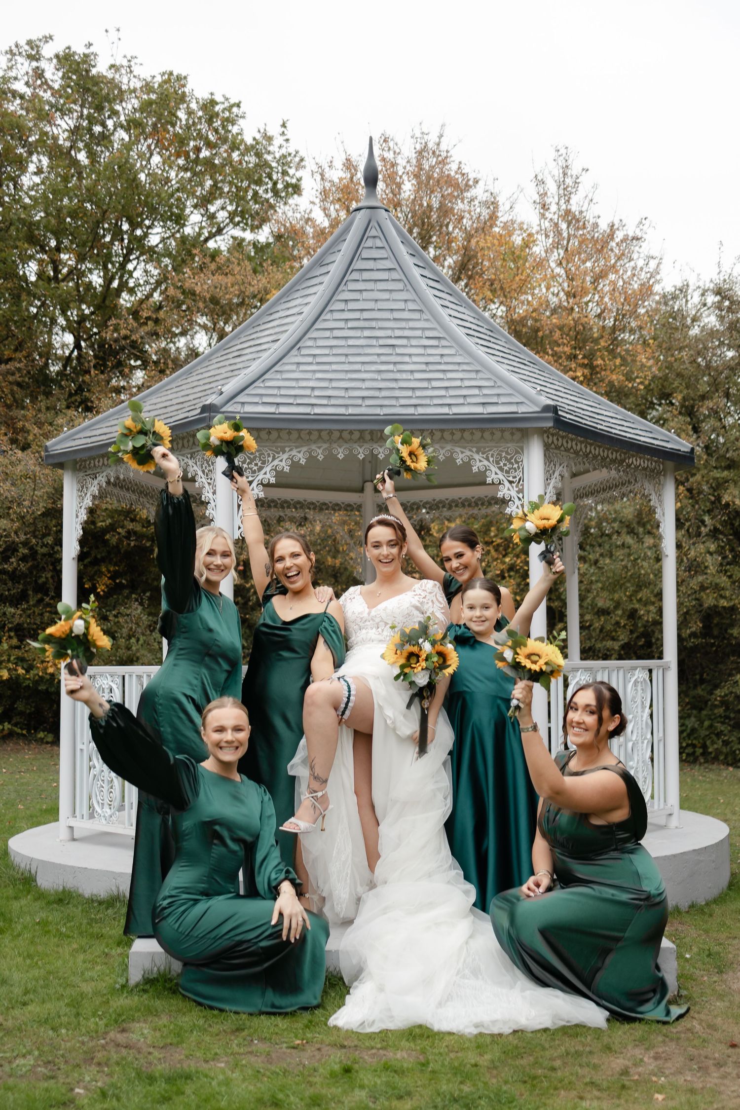 Bridal party in emerald green dresses poses playfully by white garden gazebo with sunflowers.