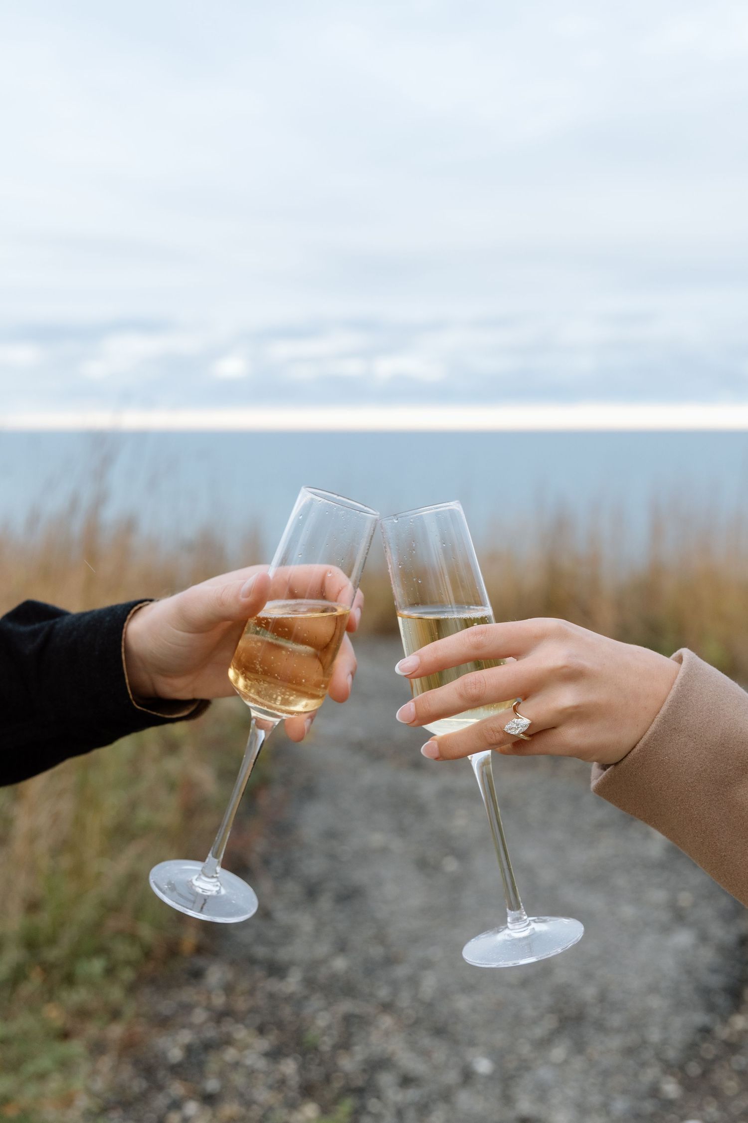 Two hands clink champagne glasses together against a blurred coastal background.