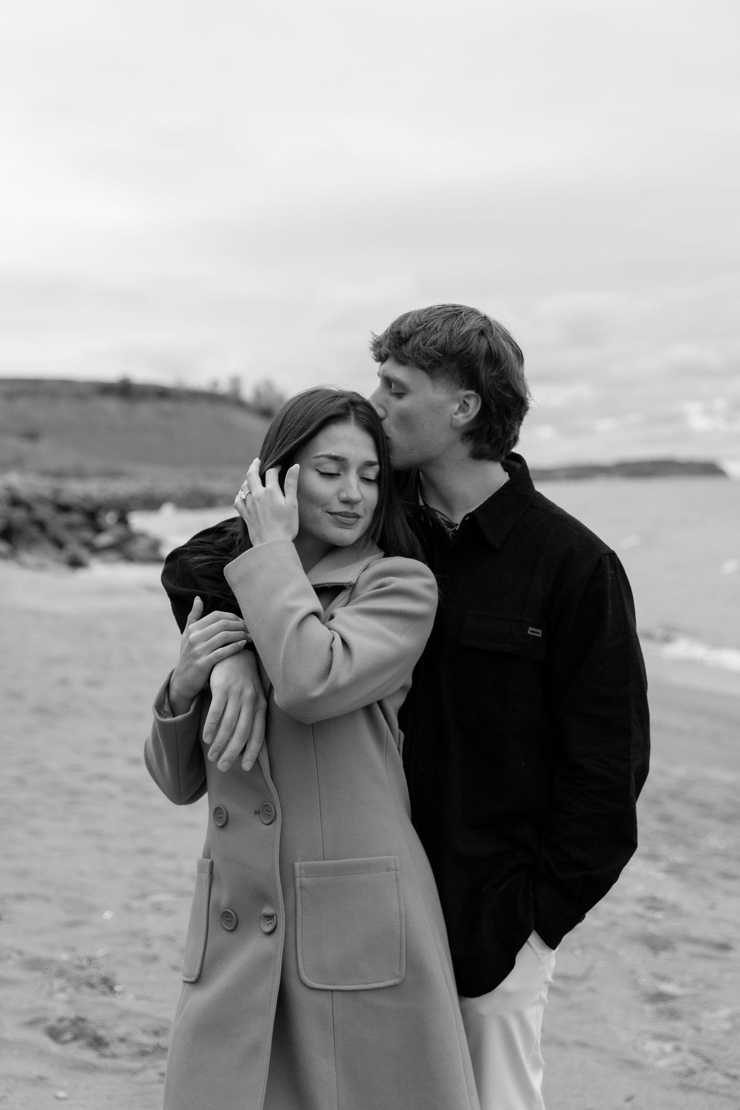 A couple shares an intimate moment together on a cloudy beach, wearing winter coats in black and beige.
