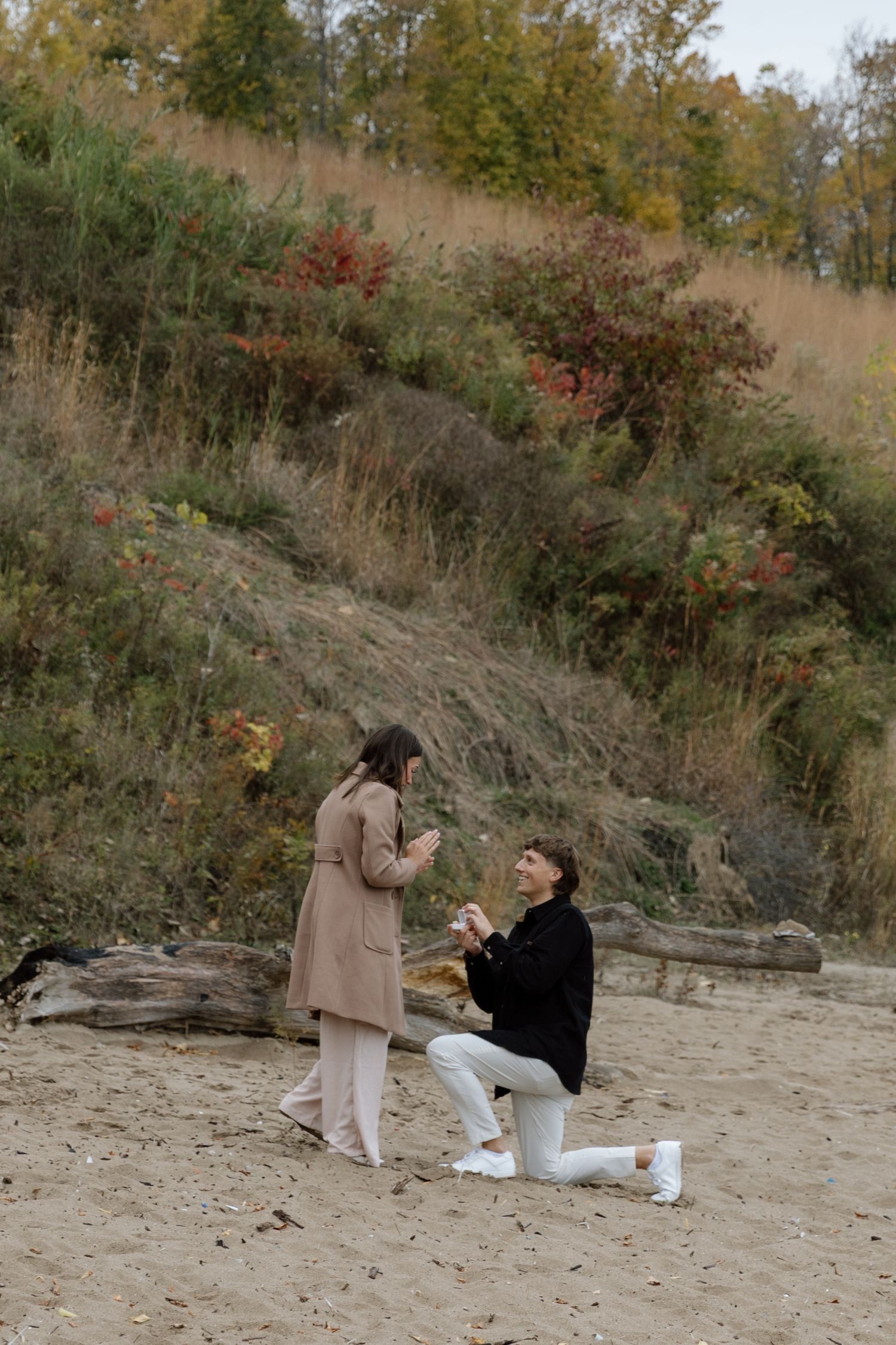 A romantic couple shares a tender moment on a sandy beach during autumn with fall foliage in the background.