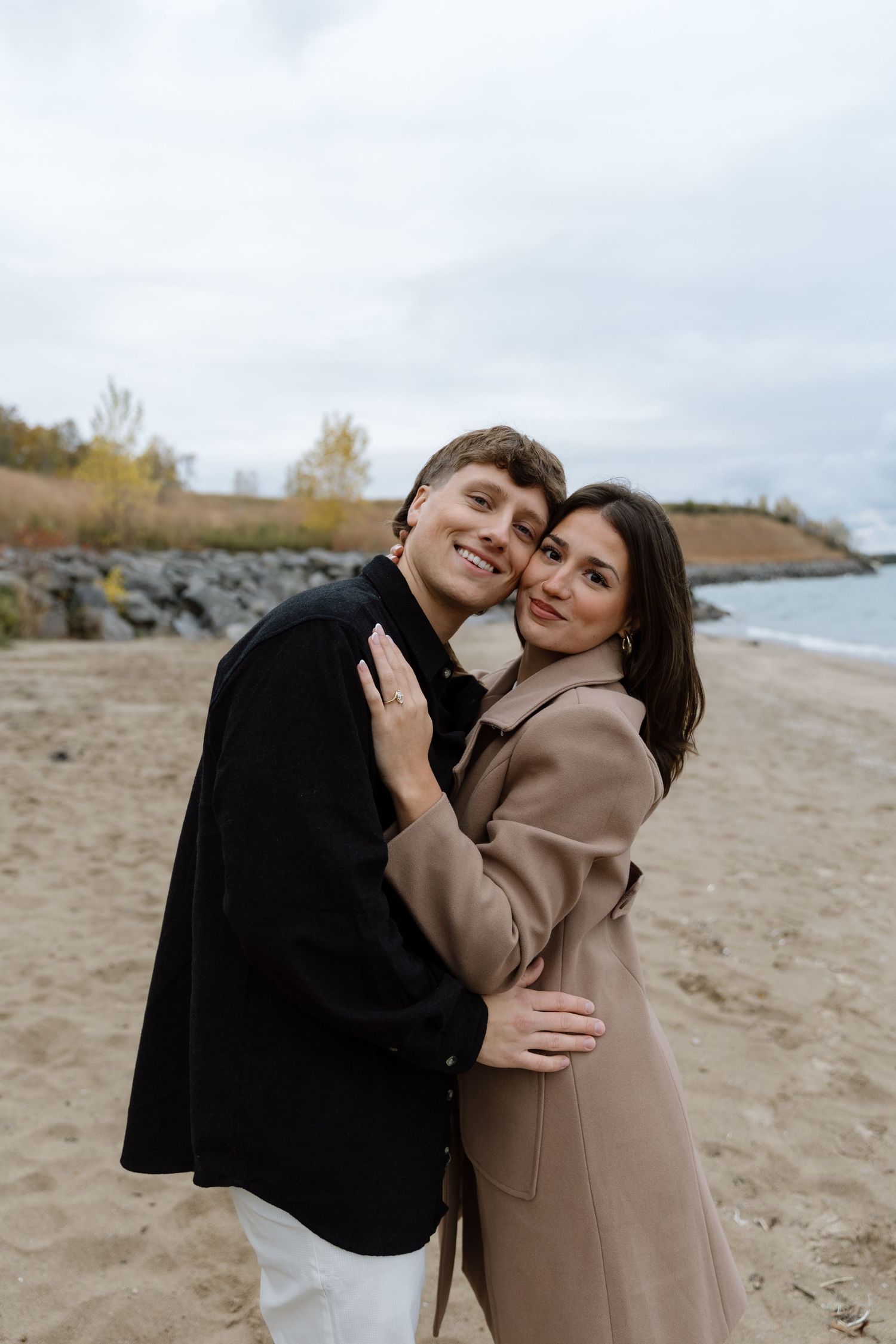 A couple in casual autumn wear shares affectionate moments on a rocky beach shoreline.