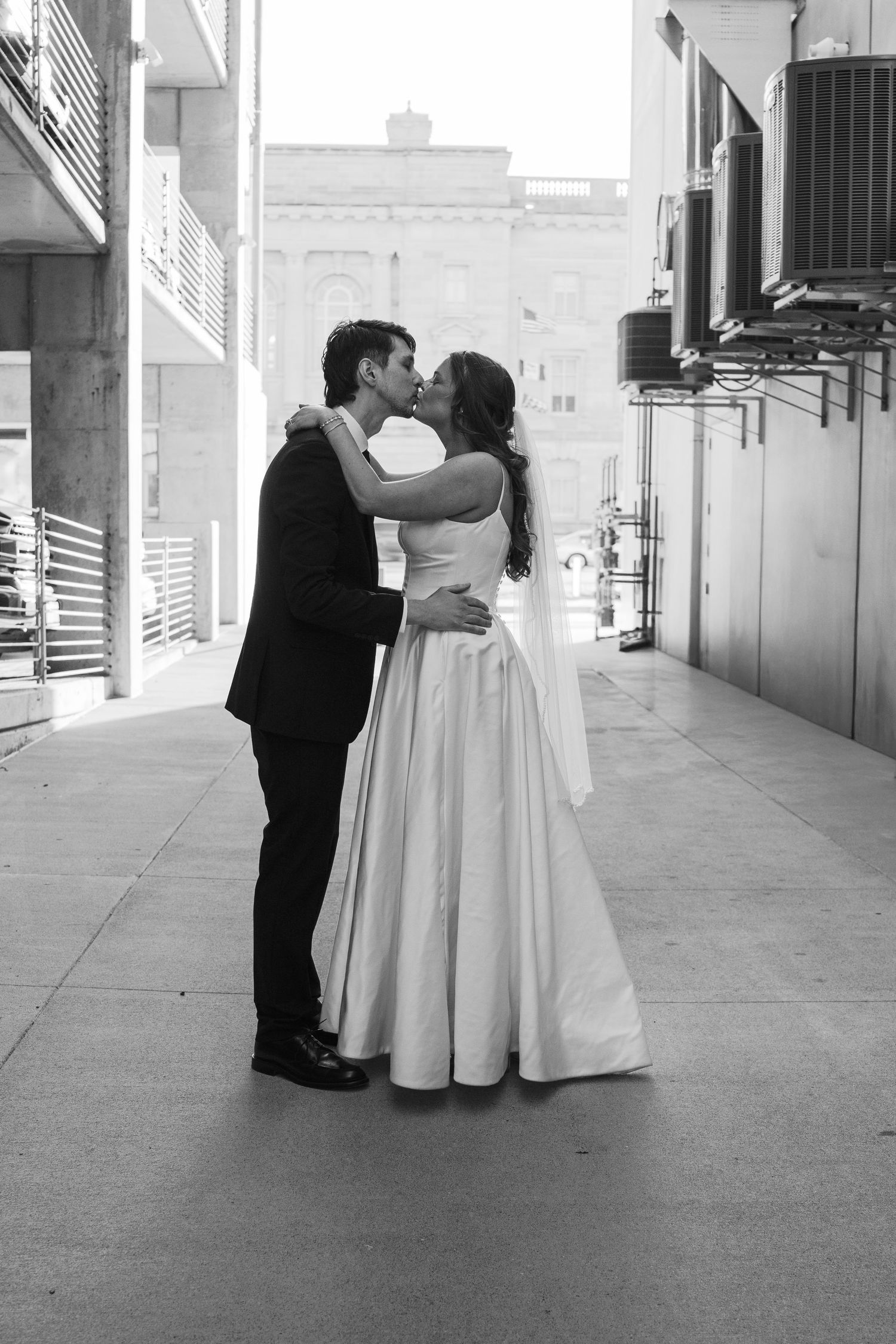 Black and white photo of a bride and groom sharing a kiss outdoors in an urban alleyway on their wedding day.