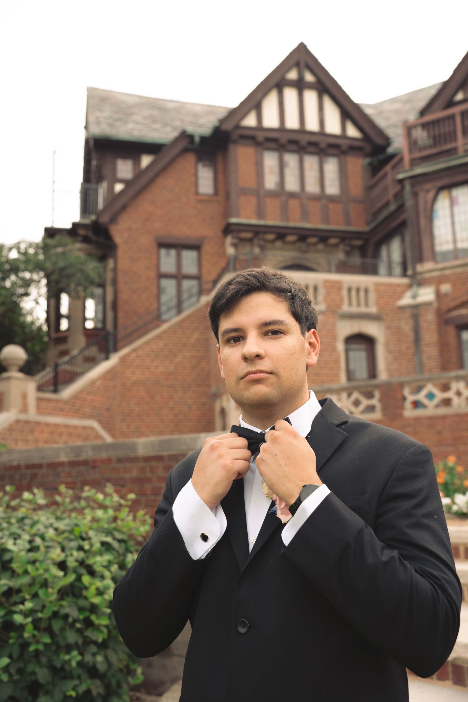 Side profile of groom in formal attire fixing his collar against brick mansion backdrop.