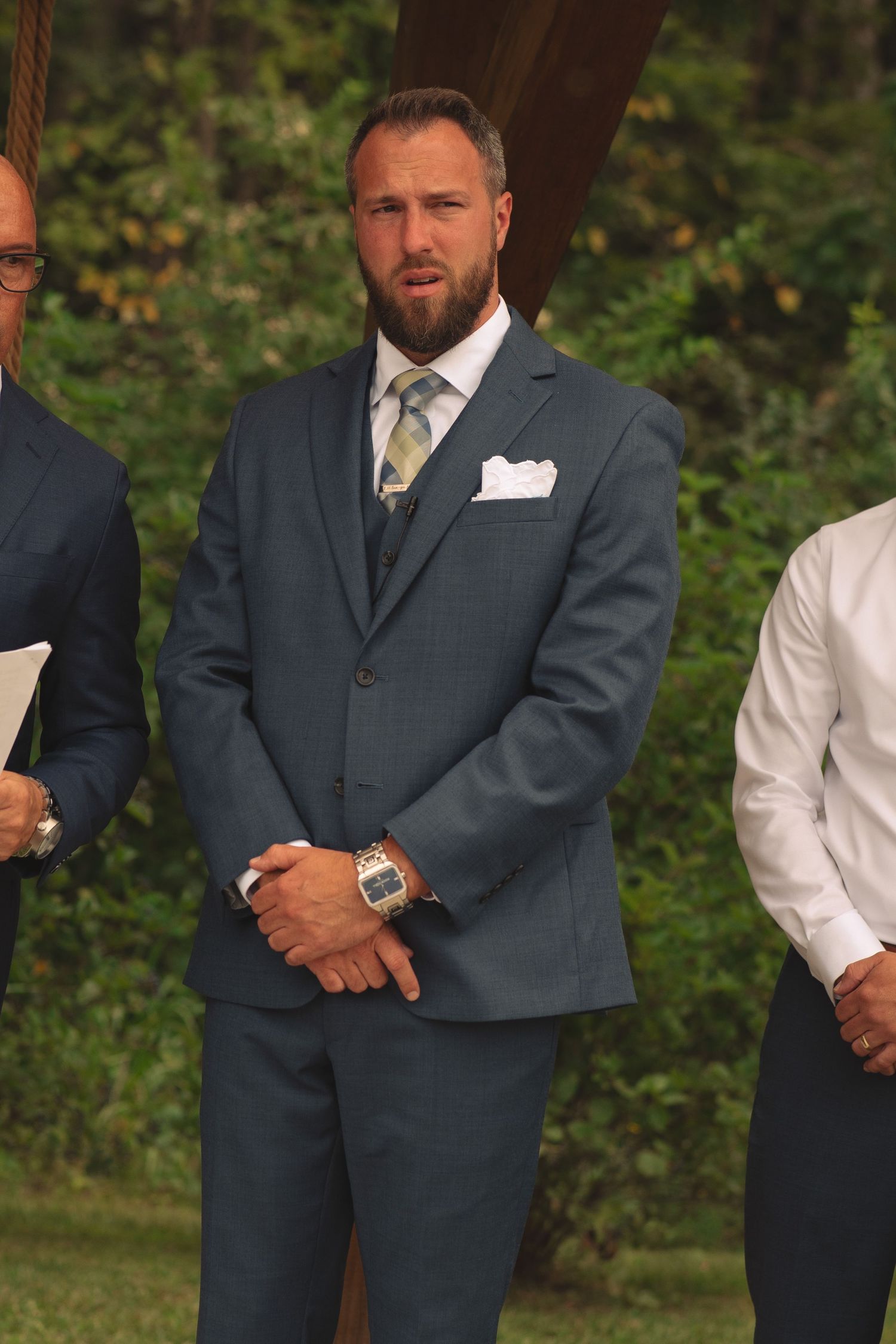 Groomsmen in charcoal suits stand at attention during outdoor wedding ceremony.