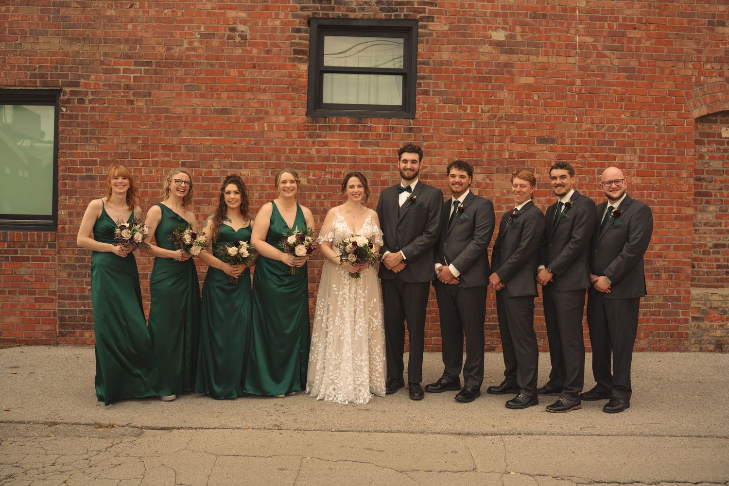 Wedding party in emerald green and black attire poses against rustic brick backdrop.