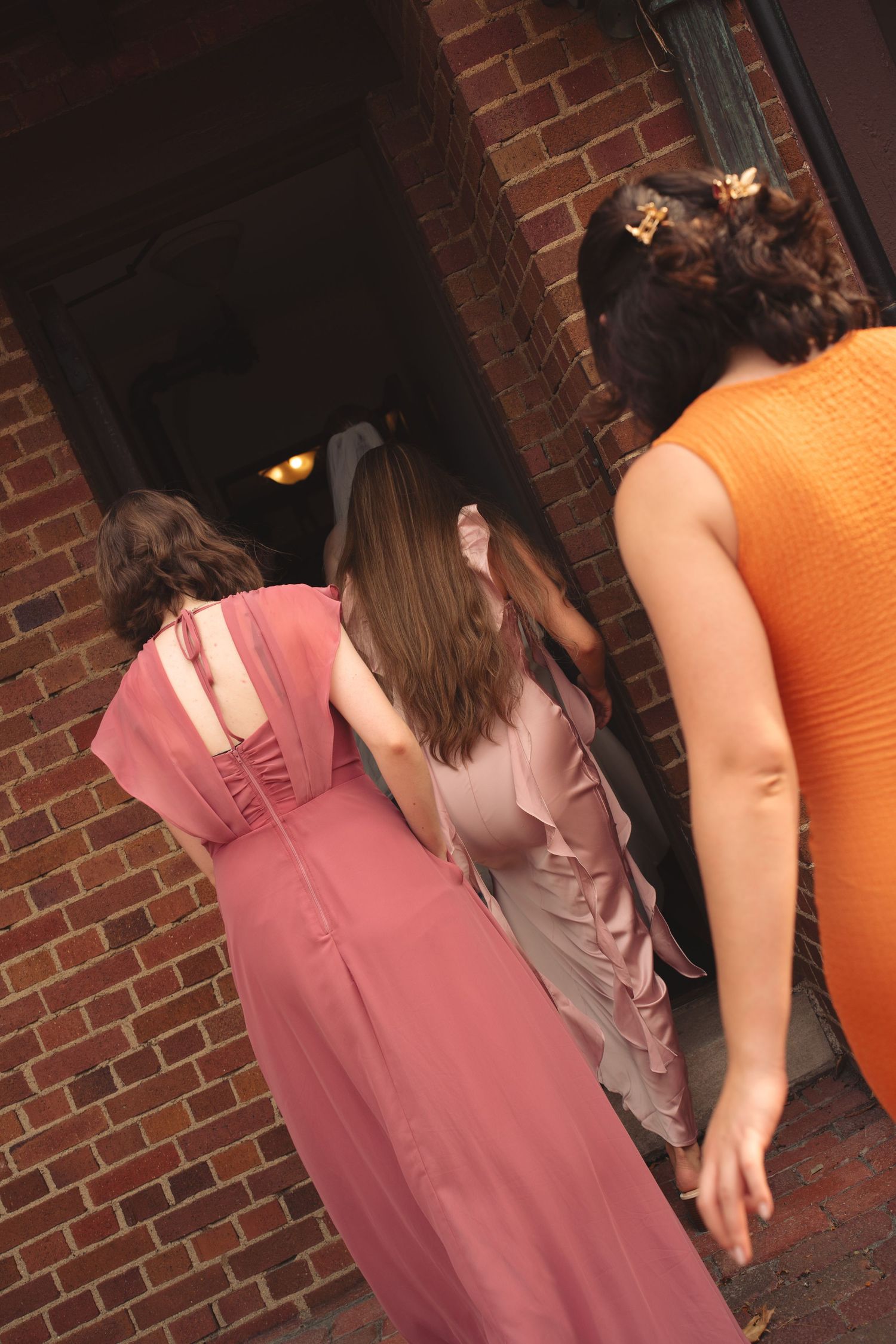 Wedding party members in pink and orange dresses stand near brick wall.