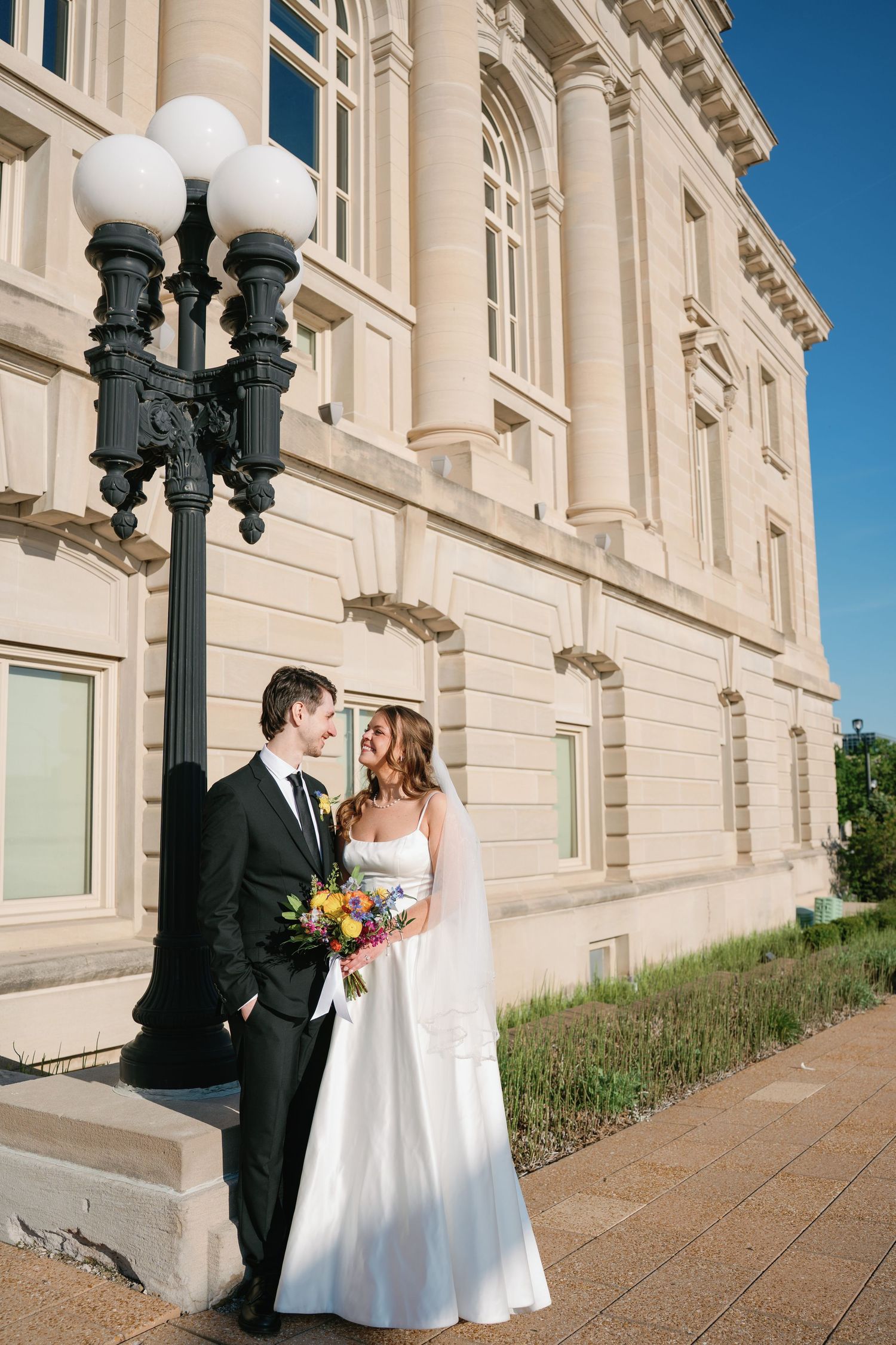 Bride and groom share a romantic moment outside a grand stone building, bride holding a colorful bouquet.