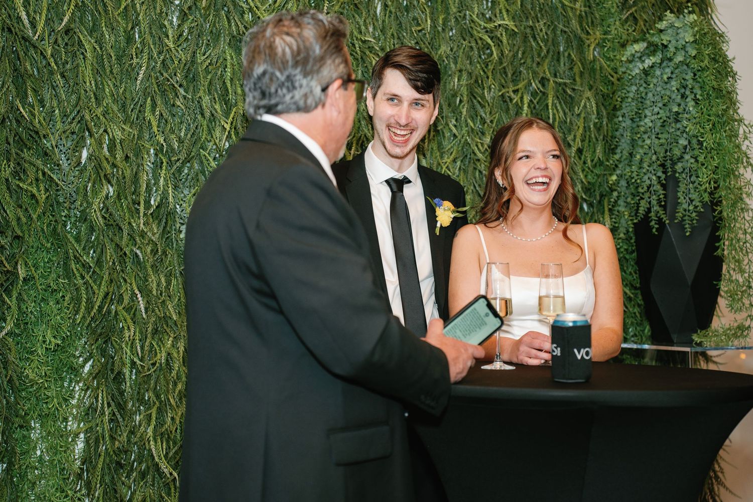 Bride and groom laughing with officiant at wedding reception near lush green ivy wall backdrop.