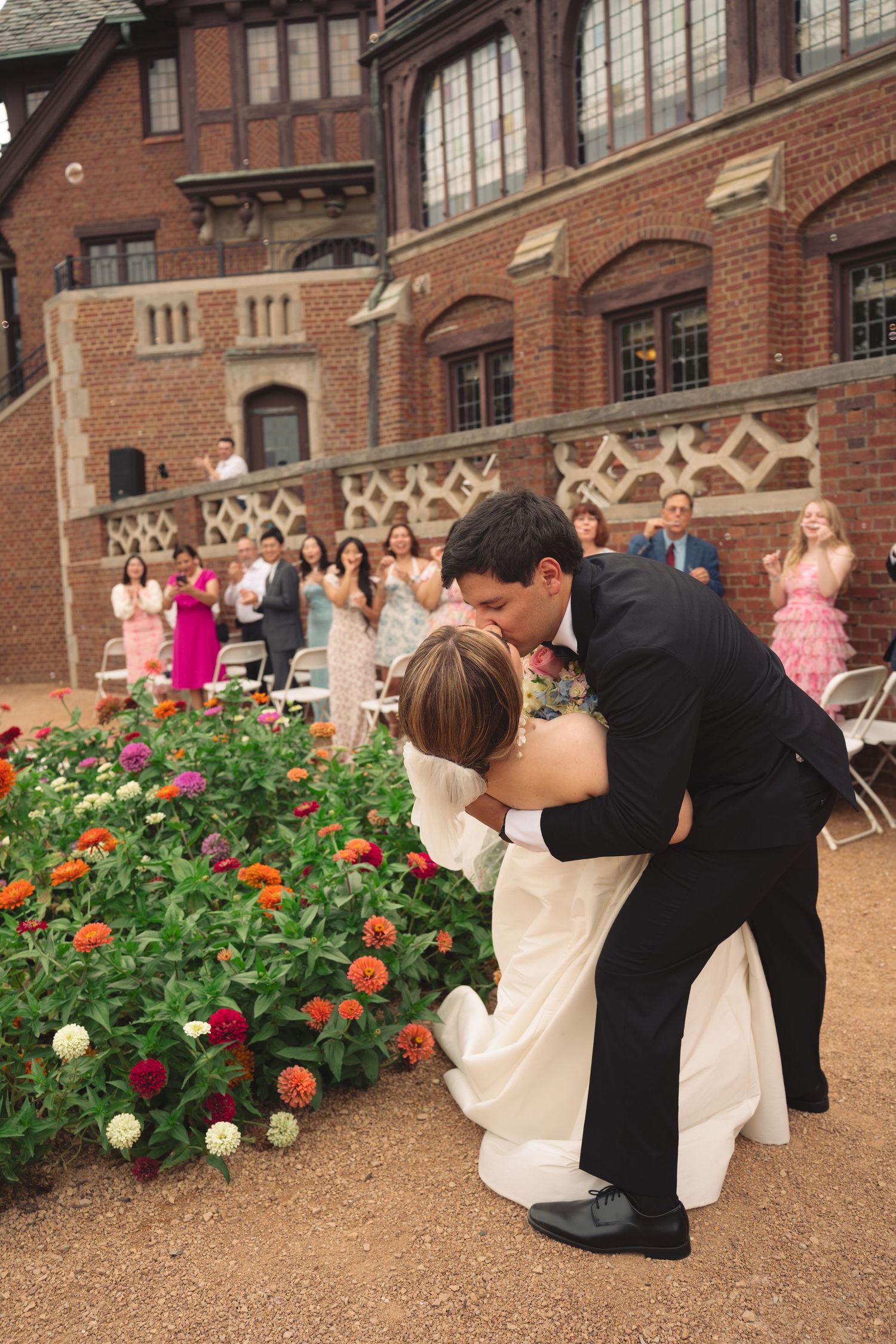 Romantic wedding dip pose in front of a historic brick building with colorful flower garden.