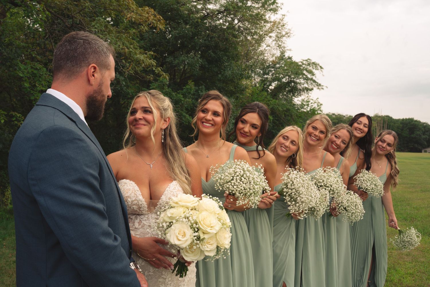 Wedding party members pose together outdoors with sage green dresses and navy suits.