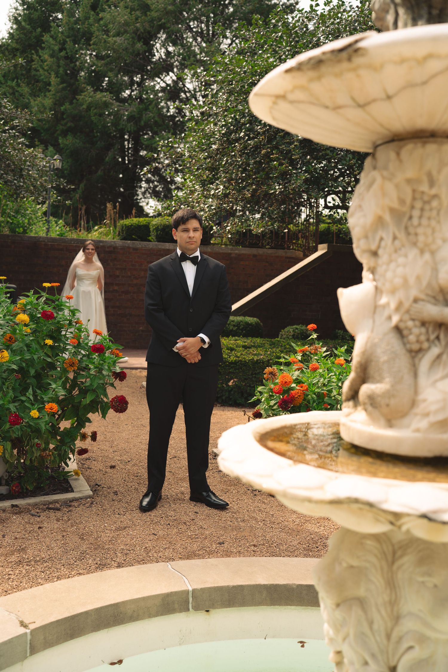 Formal wedding photo session in a garden with ornate fountain and colorful flower beds.