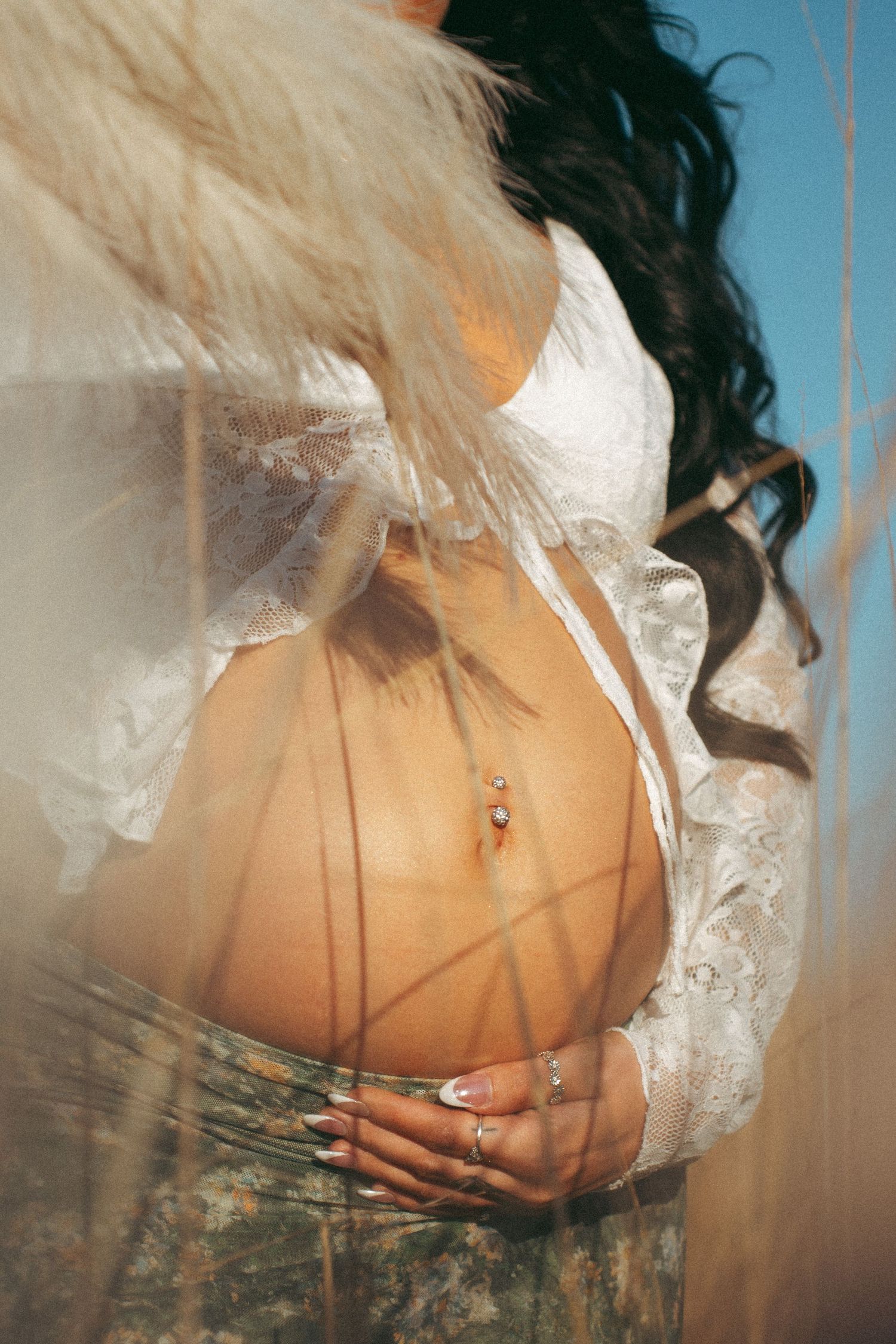 Pregnant woman in white lace top cradling baby bump, wearing belly ring and rings, with pampas grass in soft sunlight.