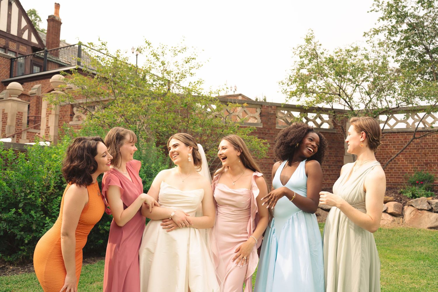 Bridesmaids in pastel dresses dance and celebrate on grass lawn near brick building.