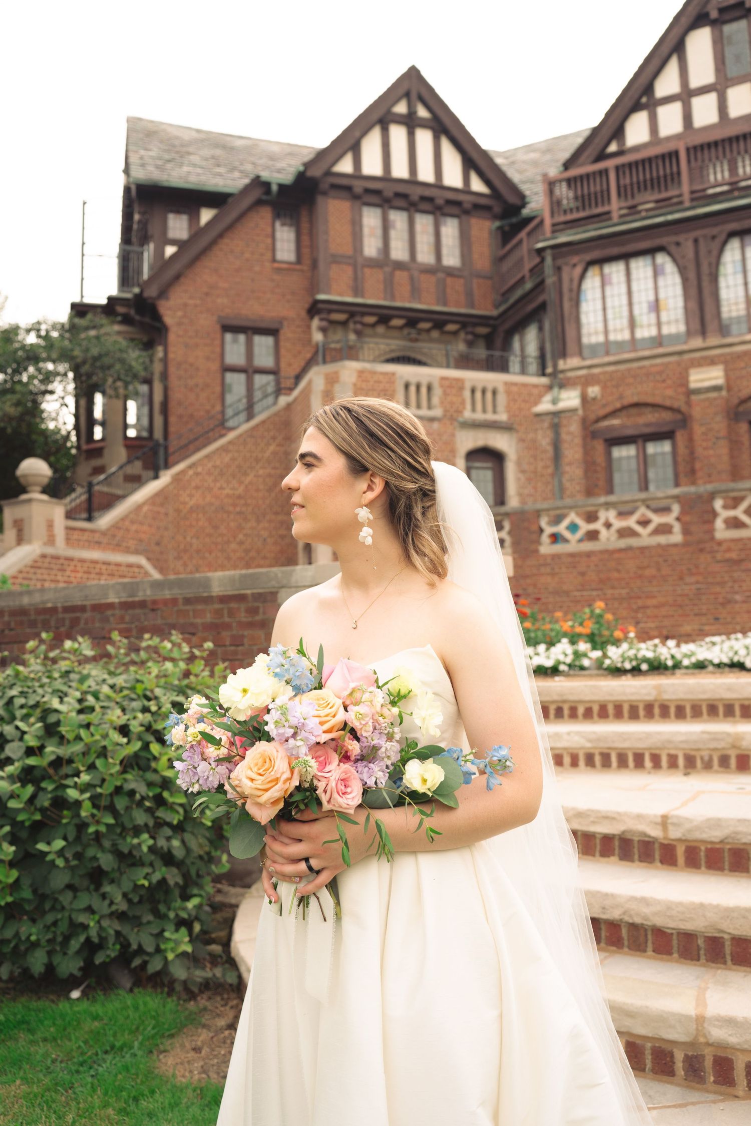 Bride in flowing white dress holds pastel bouquet on mansion steps.