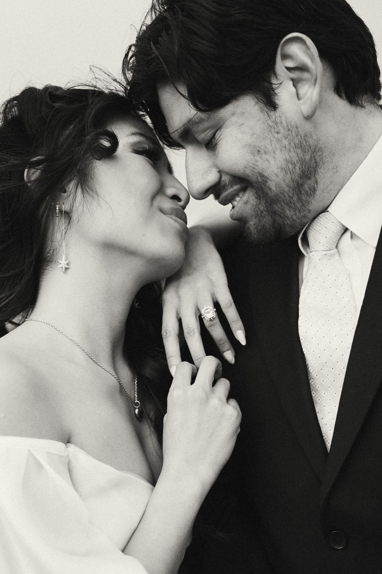 Black and white photo of a smiling couple in wedding attire, foreheads touching, woman wearing engagement ring.