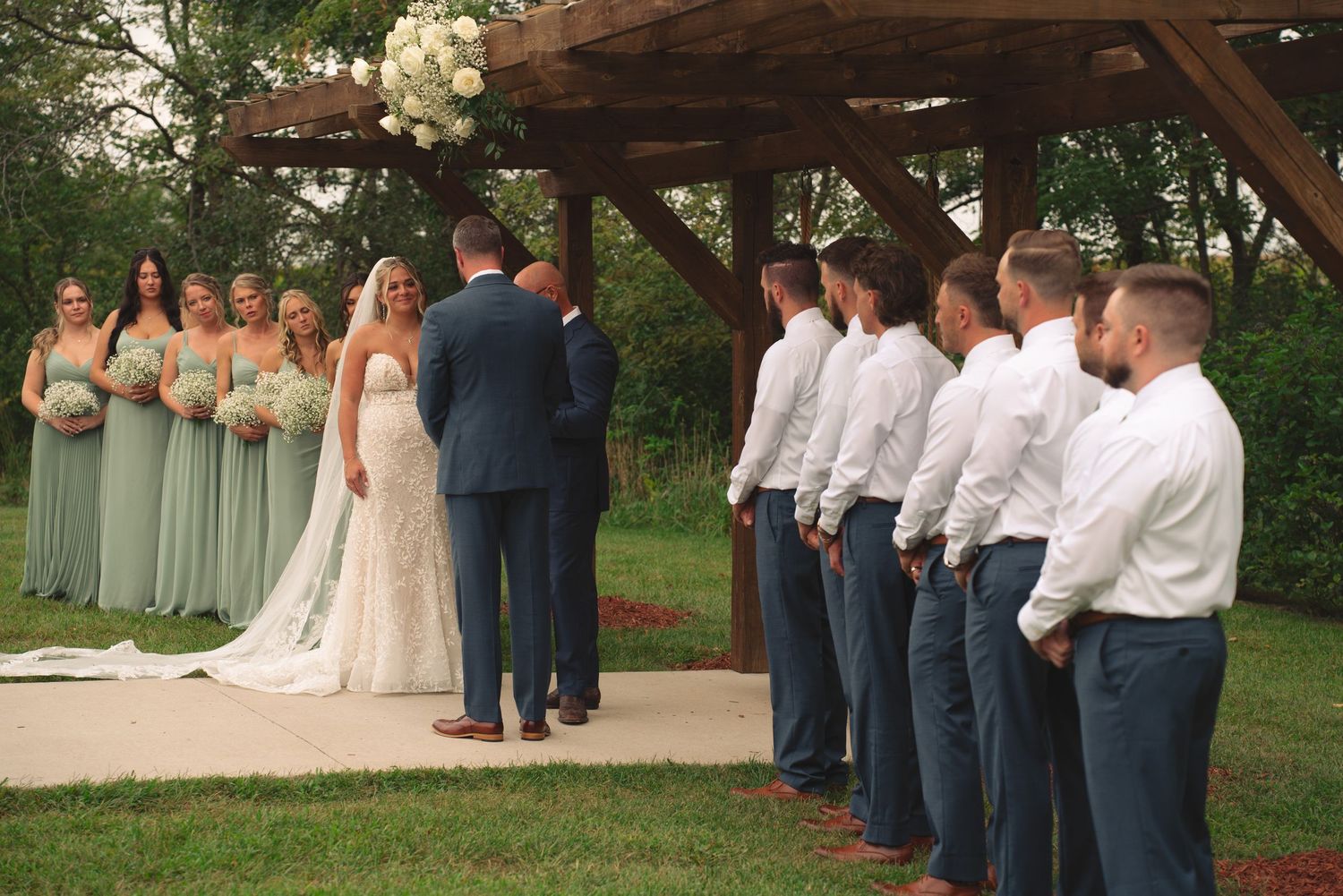 Wedding party gathers under rustic wooden pergola decorated with white floral arrangements.