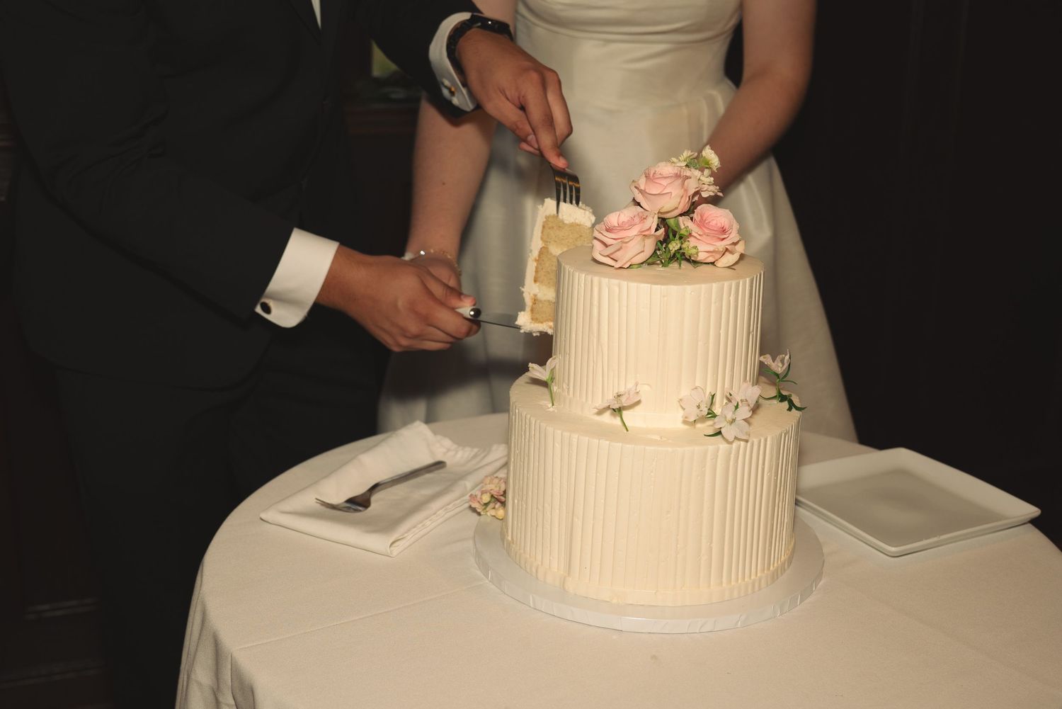Close-up of hands cutting into a white tiered wedding cake with pink floral decorations.