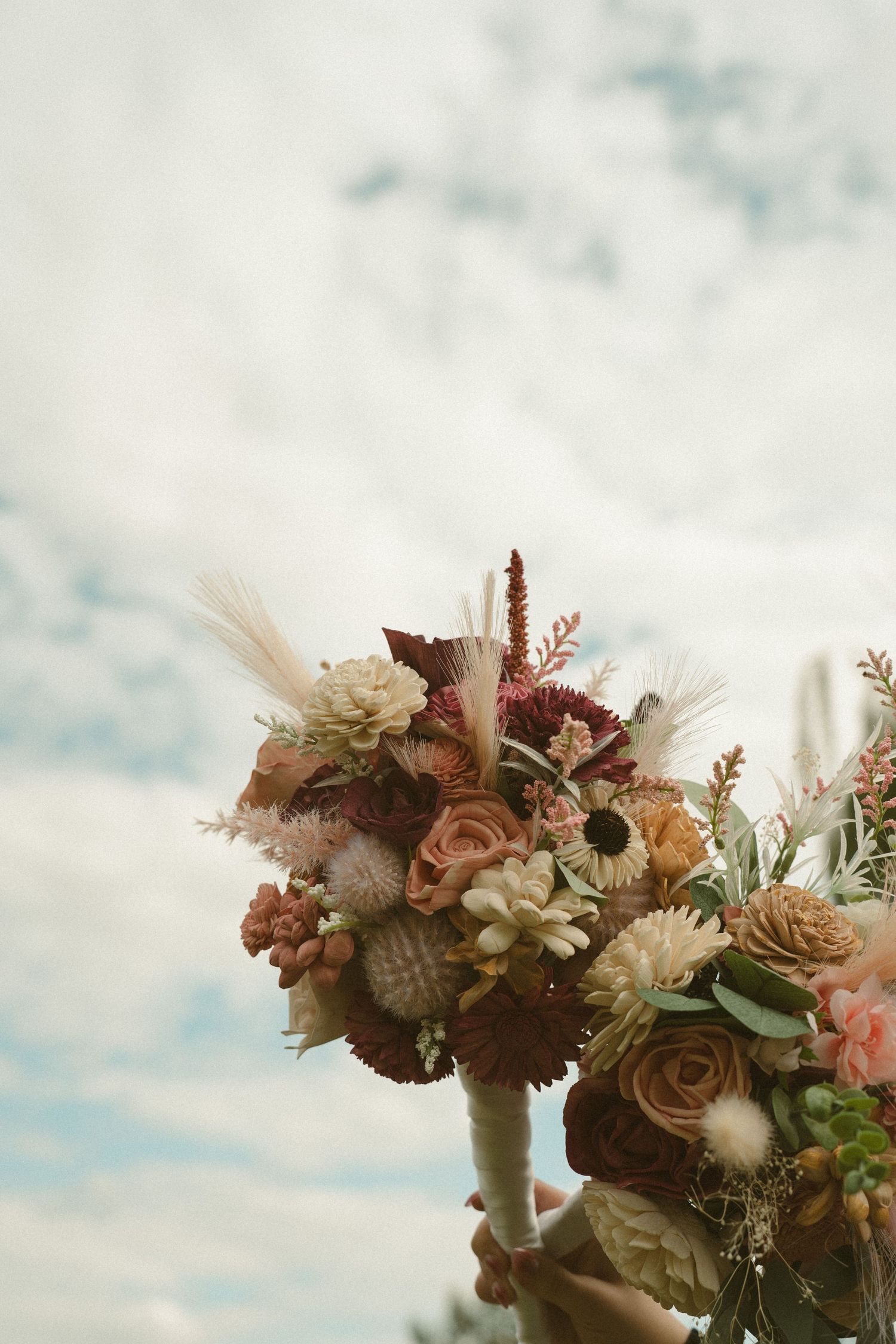 Rustic wedding bouquet with dried flowers and pampas grass against cloudy sky.