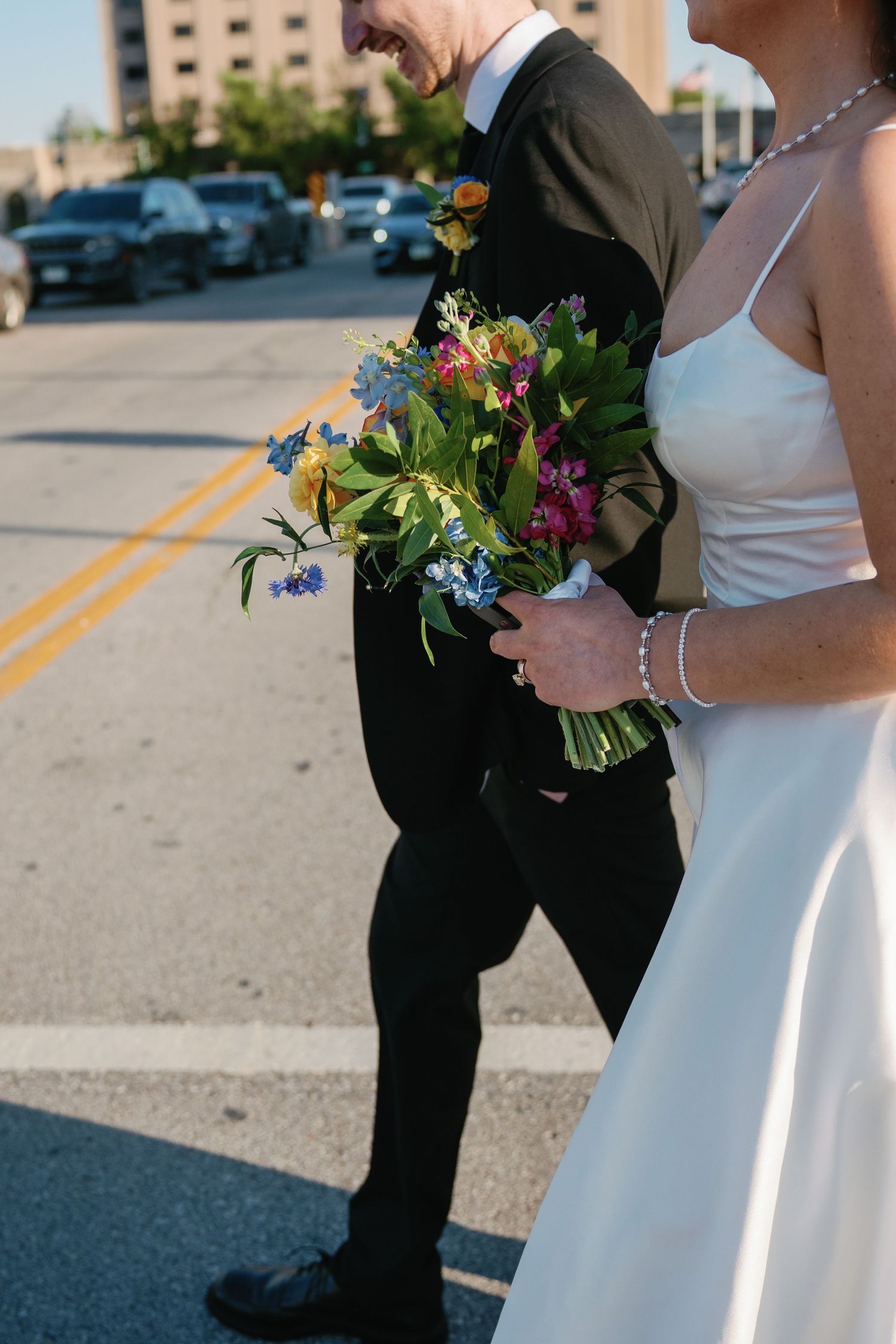 Bride holding colorful wildflower bouquet walks with groom in black suit across urban street on wedding day.