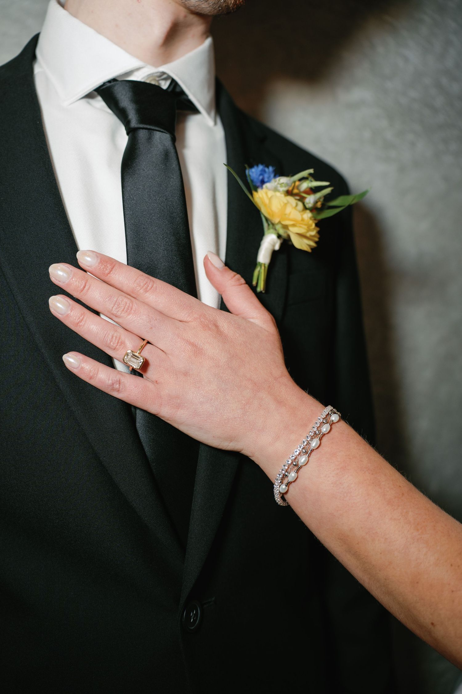 Bride's hand with diamond ring and bracelet resting on groom's black tuxedo with floral boutonniere.
