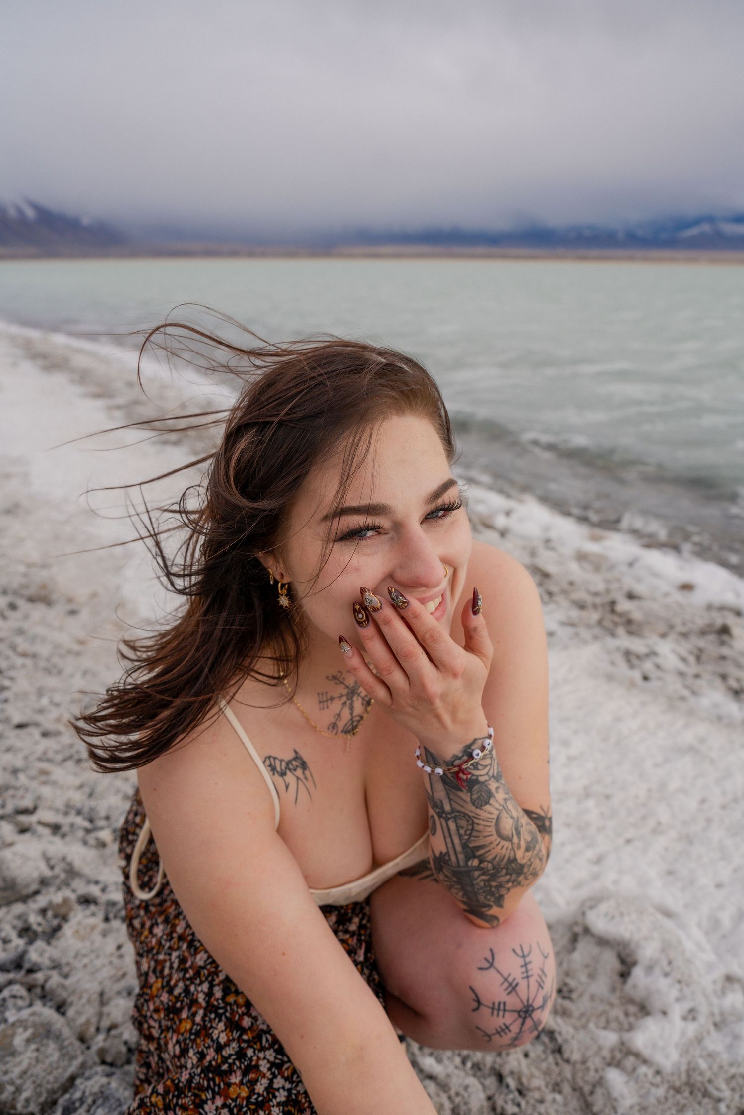 Person with windswept hair poses on snowy shoreline with mountains in background.